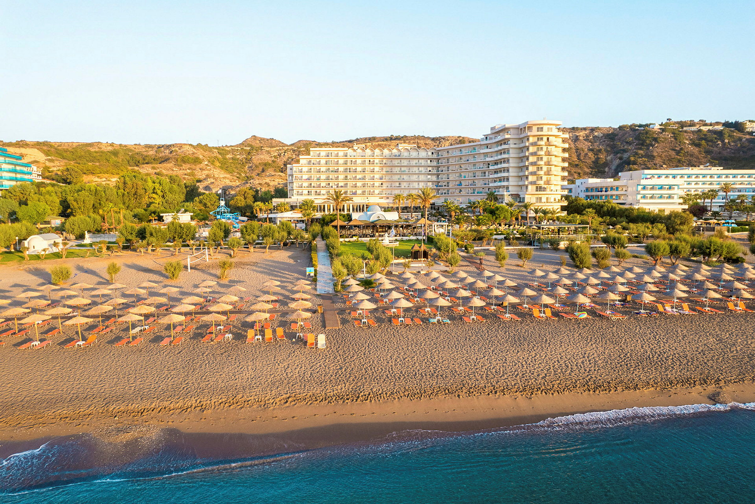 Strand med parasoller og solsenger, hotell og grønne områder i bakgrunnen. (AI-generert tekst)