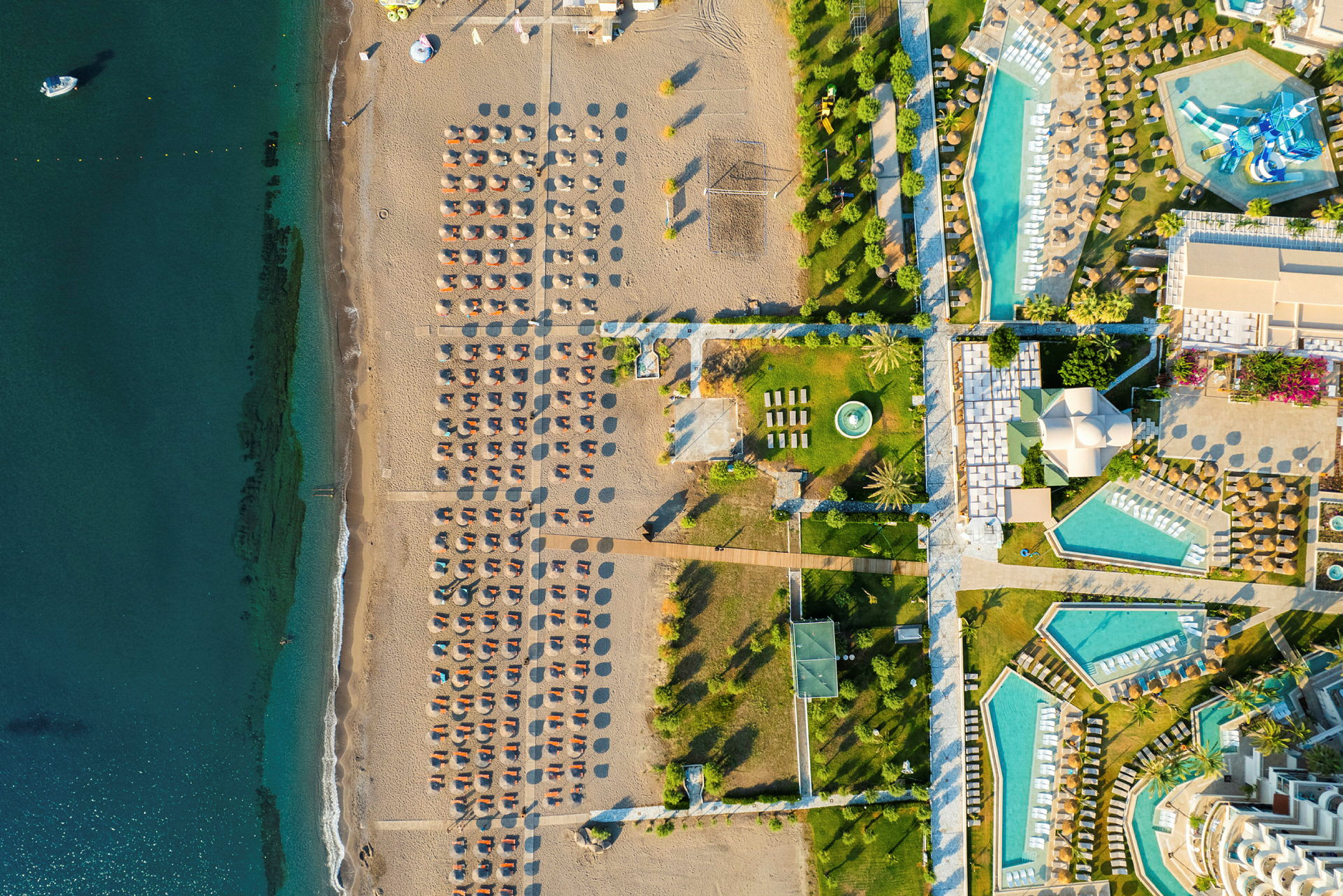 Luftfoto av en strand med solsenger og parasoller ved siden av et hotellområde med bassenger og grøntområder. (AI-generert tekst)