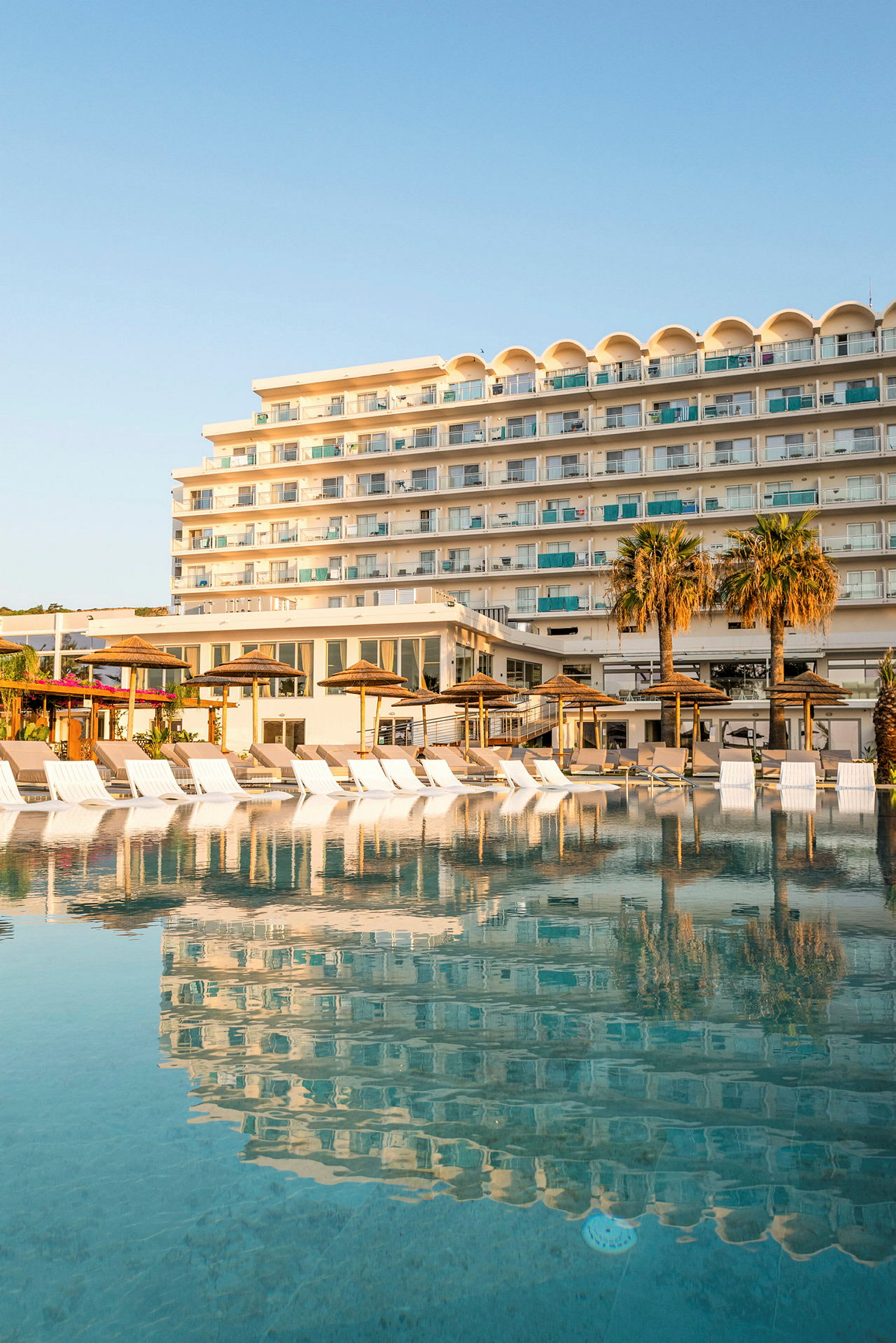 Hotellbygning ved et stort basseng med solsenger og parasoller i forgrunnen. (AI-generert tekst)