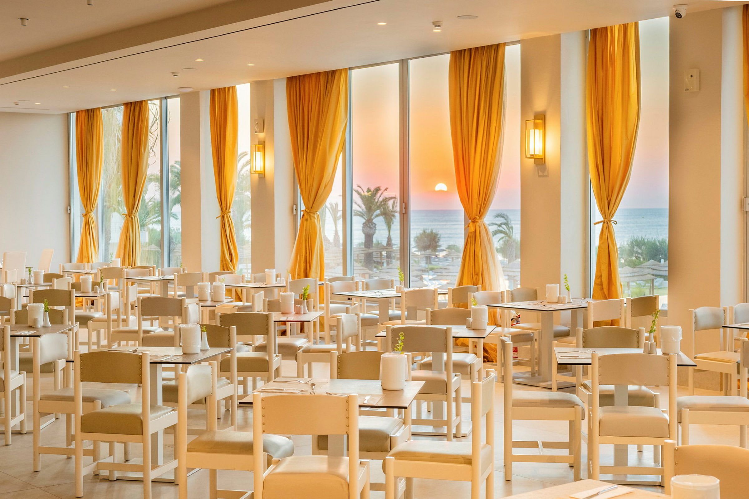 En restaurant med bordoppsett og hvite stoler. Store vinduer med gule gardiner viser en solnedgang over havet og palmer. (AI-generert tekst)