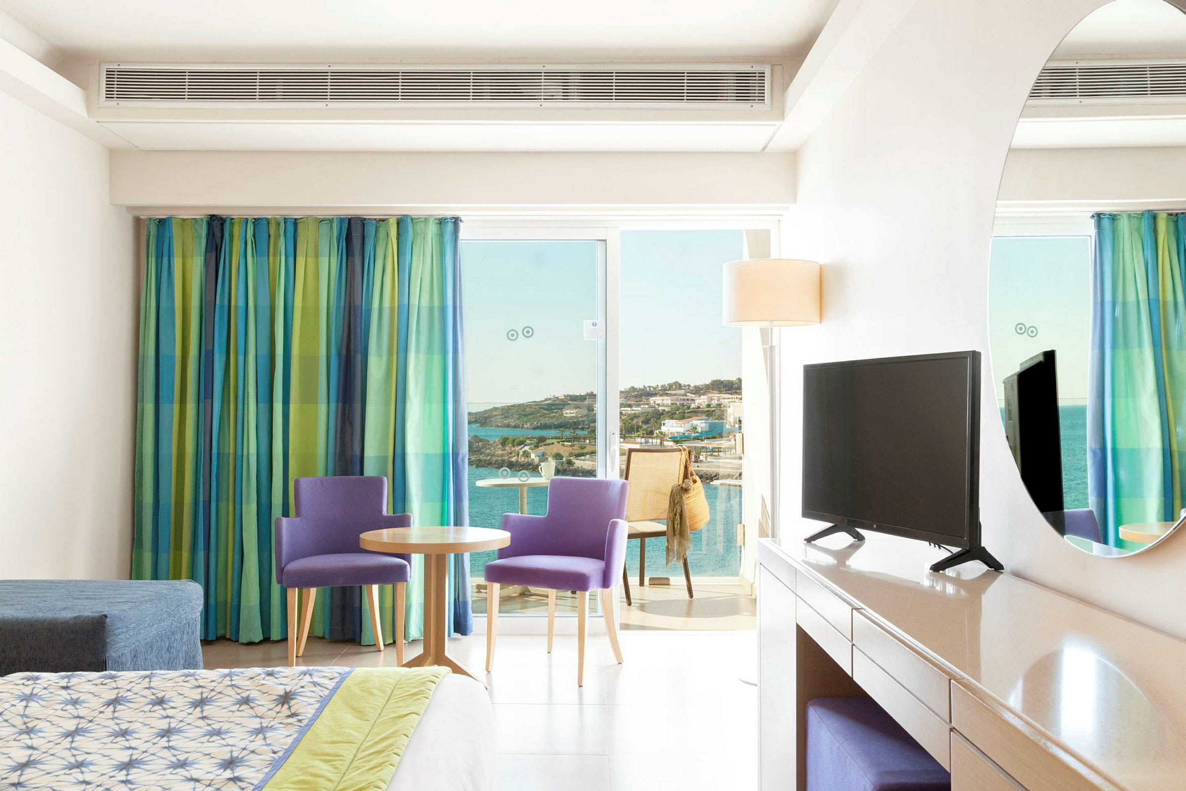 Et hotellrom med seng, TV på kommode og sittegruppe foran en glassdør som fører til balkong med utsikt over havet. (AI-generert tekst)