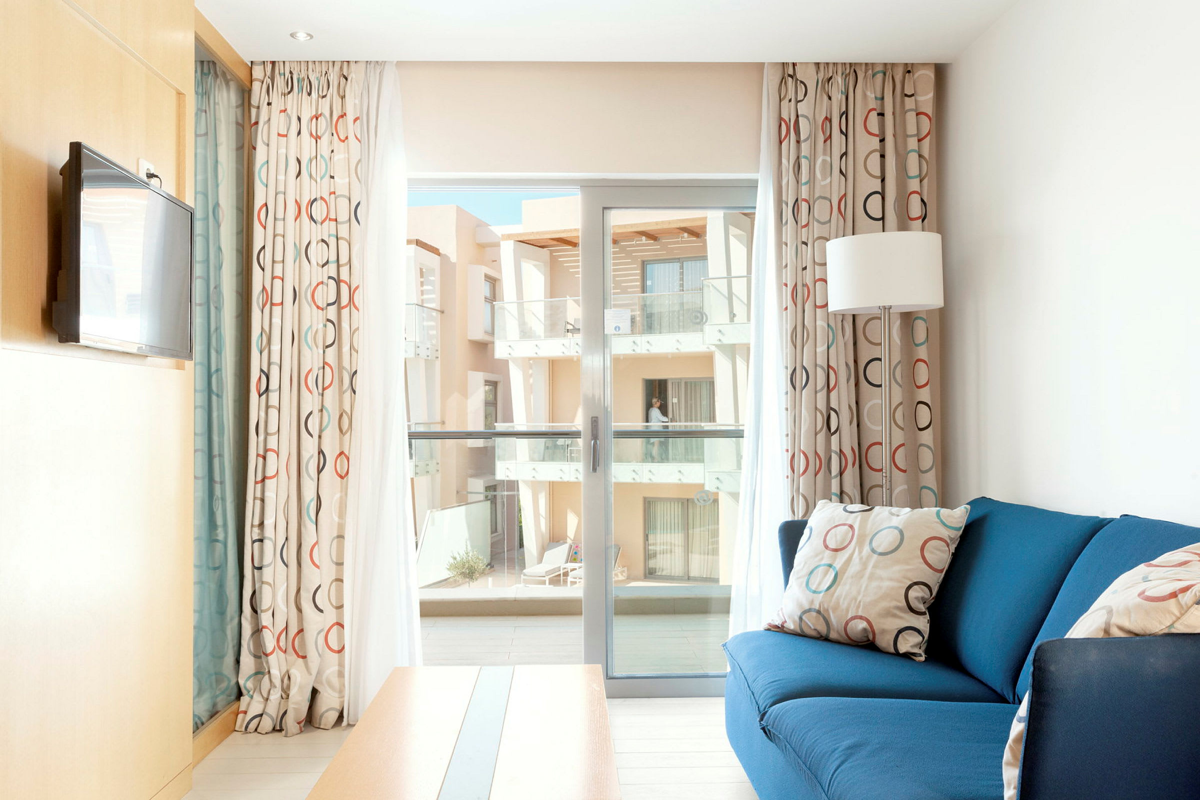 Et lyst hotellrom med blå sofa, gardiner med sirkelmønster og utgang til en balkong. (AI-generert tekst)