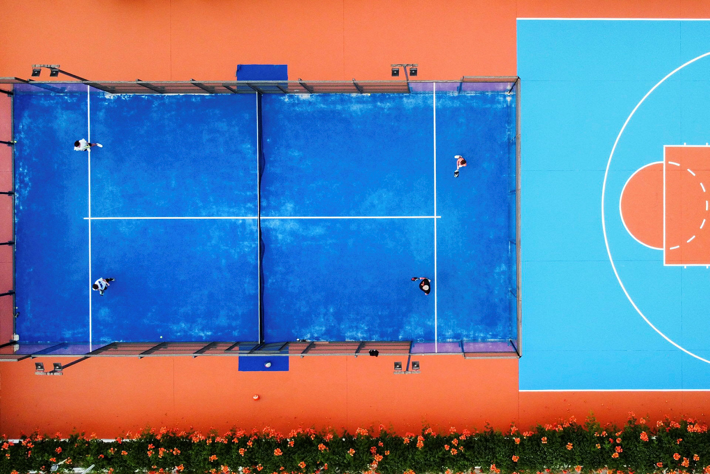Luftbilde av en blå padelbane med personer som spiller, ved siden av en oransje og blå basketballbane. (AI-generert tekst)