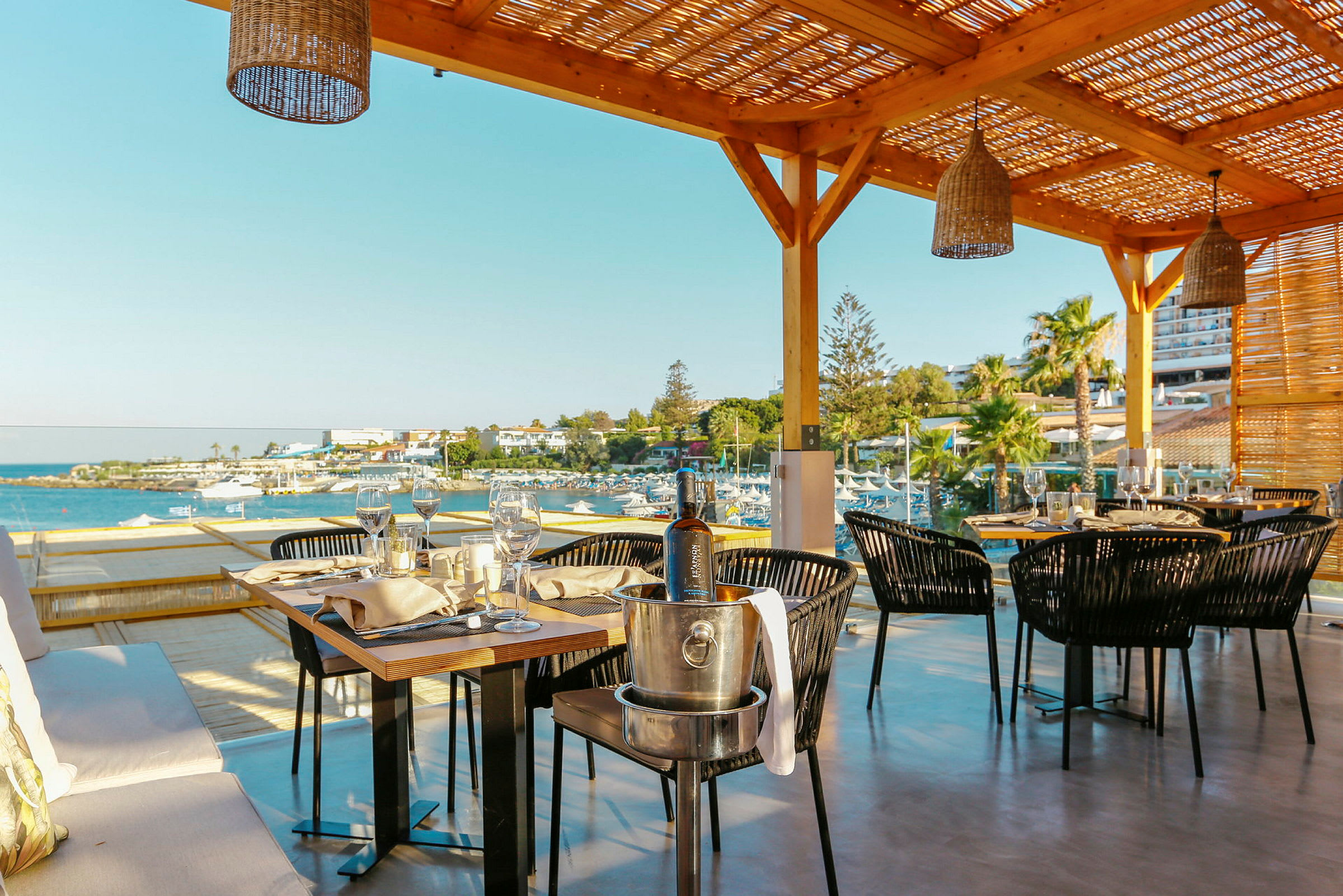 Utendørs serveringsområde med bord og stoler under en pergola, med utsikt over sjø og strand. (AI-generert tekst)