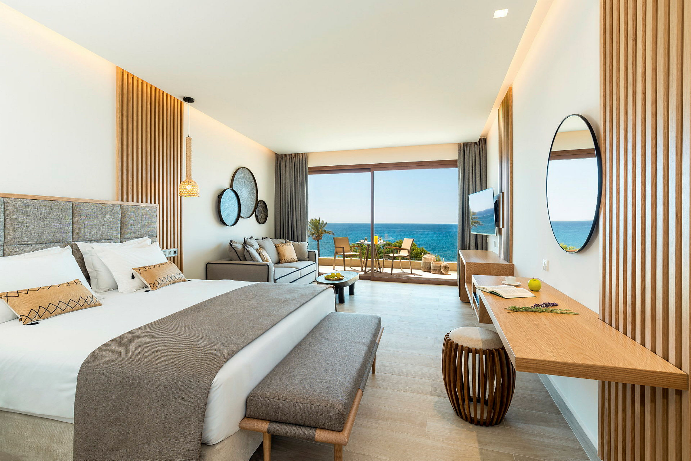 Et stilrent hotellrom med en dobbeltseng, sofa og et skrivebord. Store glassdører åpner seg mot en balkong med utsikt over havet. (AI-generert tekst)