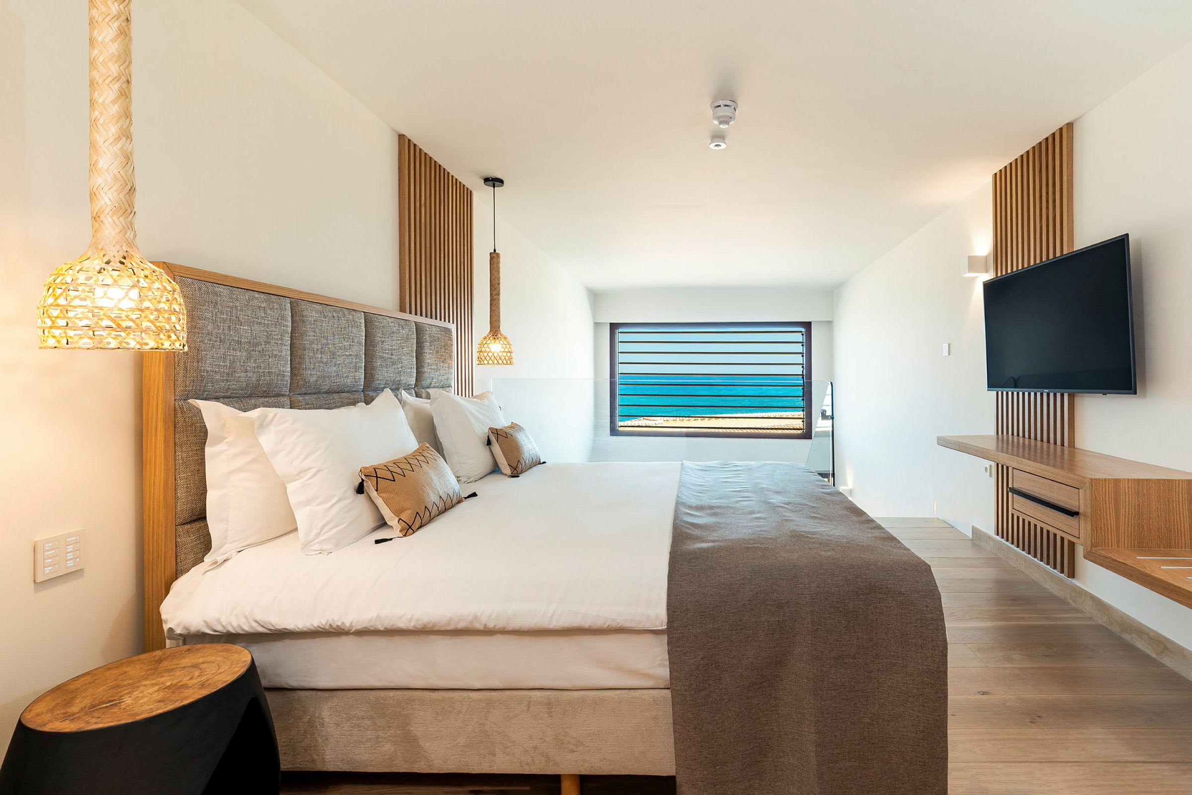 Hotellrom med seng, lamper over sengen og en veggmontert TV. Et vindu med havutsikt er i bakgrunnen. (AI-generert tekst)