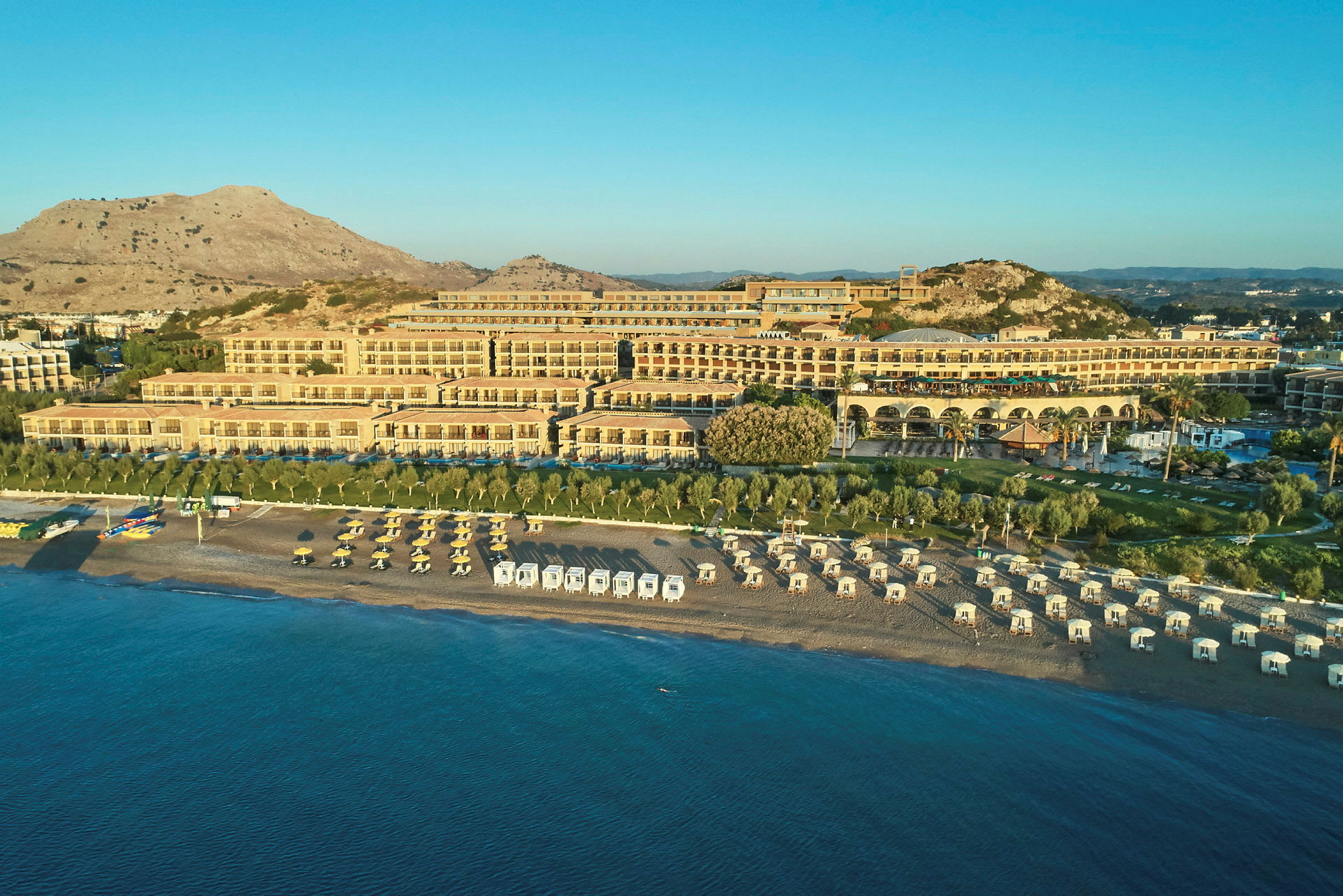 Ved en solrik kyst ligger et stort hotell med flere terrasser og rekker med solsenger og parasoller på stranden. (AI-generert tekst)