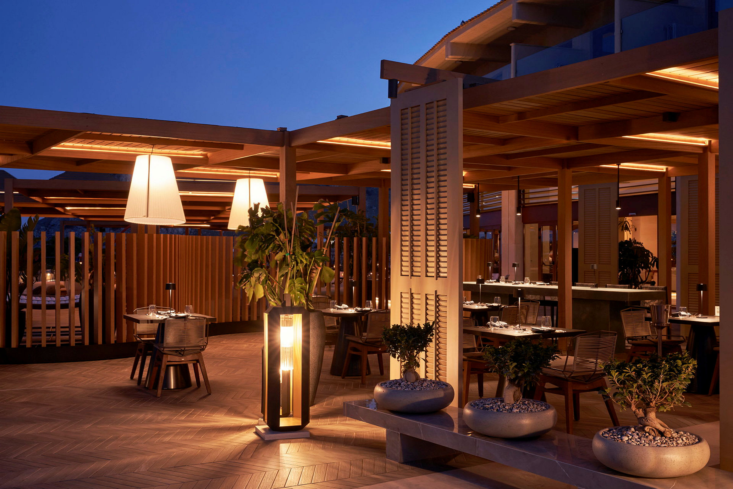 Utendørs serveringsområde med bord dekket til middag, omgitt av stilren arkitektur og planter, under kveldsbelysning. (AI-generert tekst)