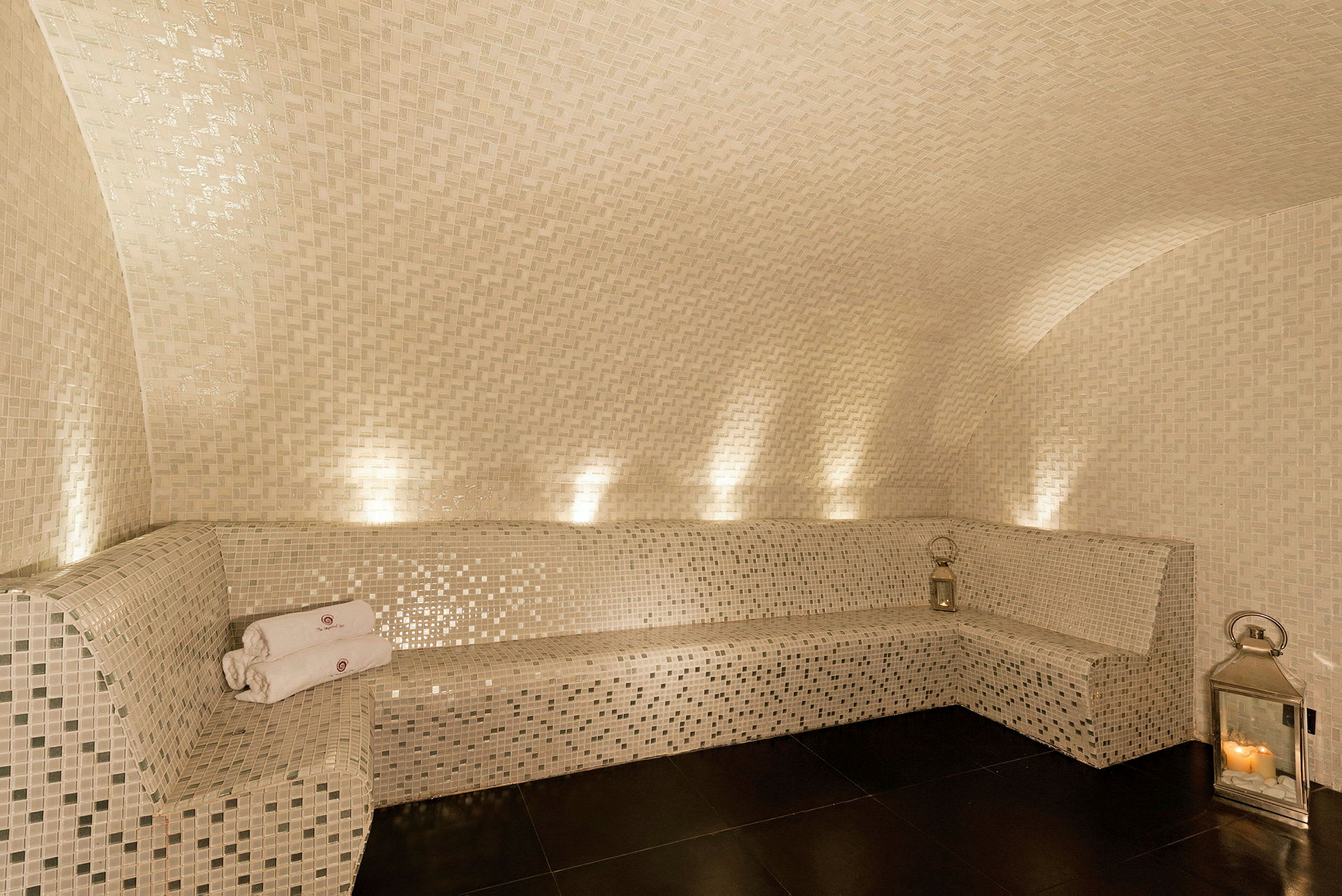 Mosaikkledde vegger og benk med lyslykt og sammenbrettede håndklær i et elegant spaområde. (AI-generert tekst)