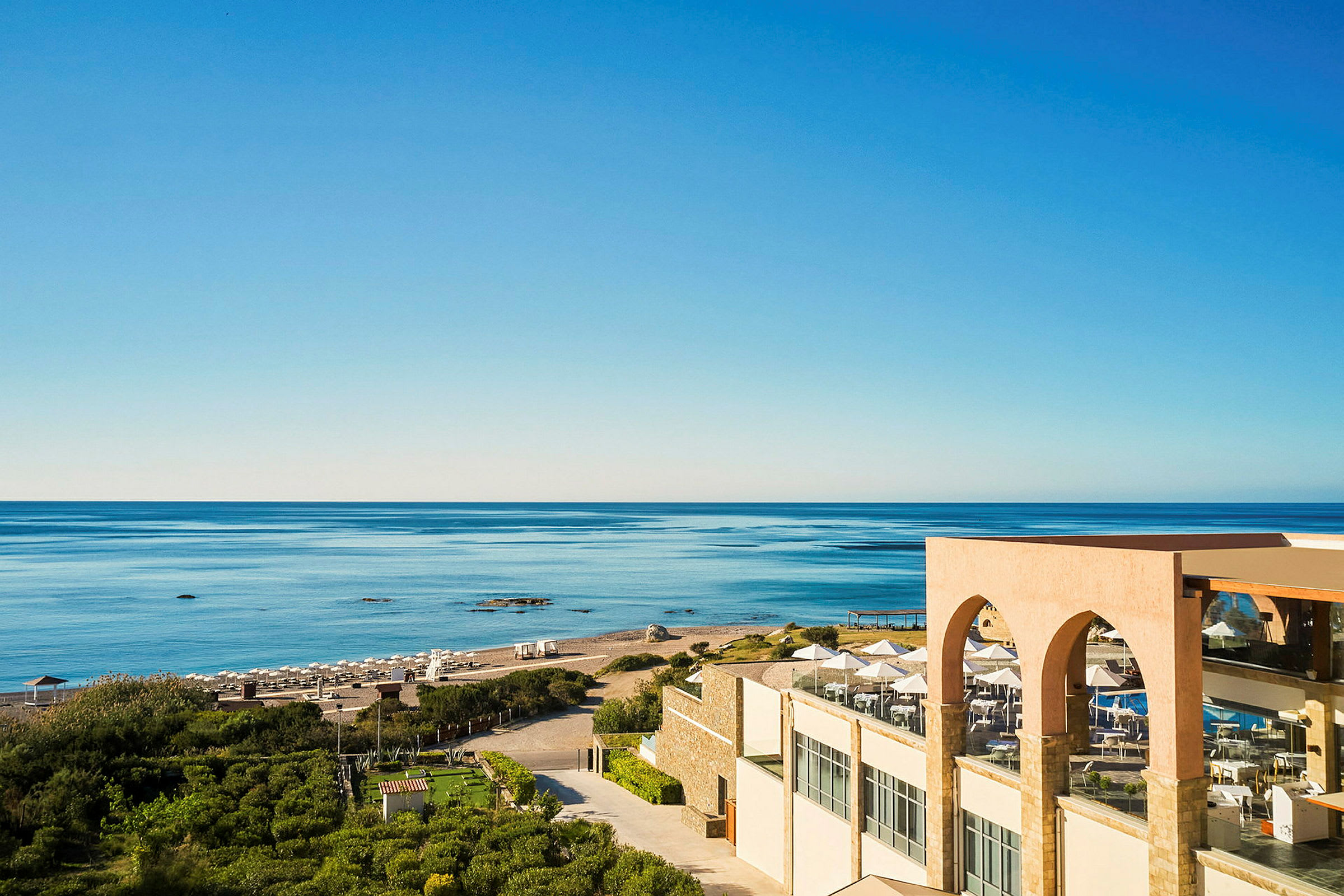 Utsikt over en sandstrand, havet og et hotell med terrasse i forgrunnen. (AI-generert tekst)