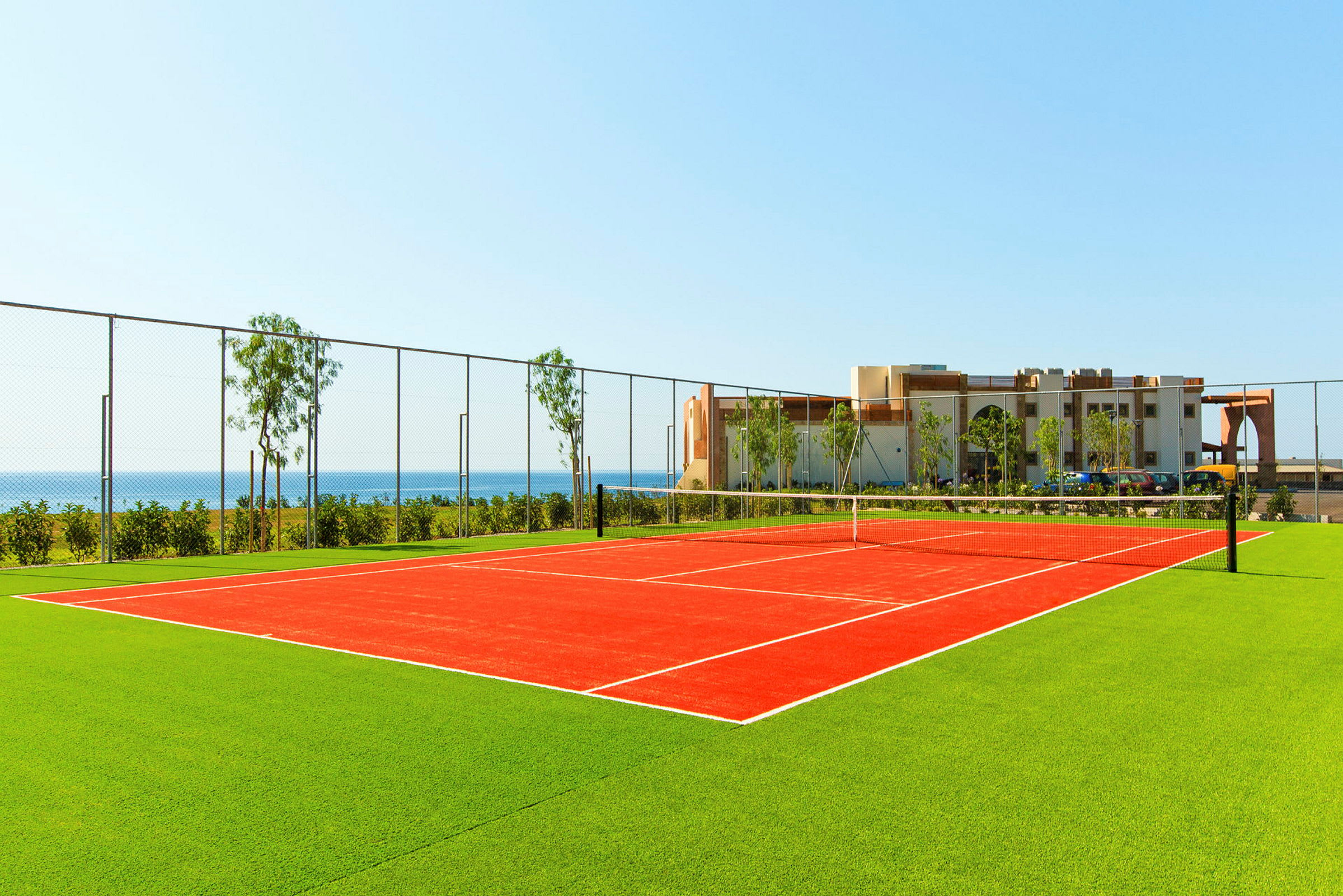En tennisbane med rød belegg omgitt av grønt gress, med havet og en bygning i bakgrunnen. (AI-generert tekst)