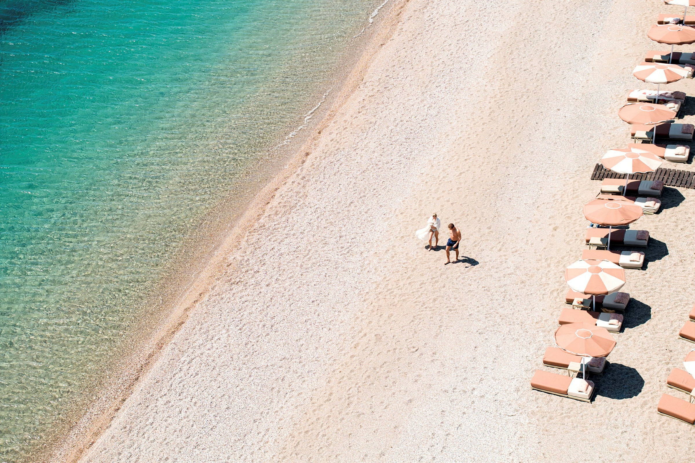 En strand med klart vann og parasoller langs den ene siden. To personer går på sanden. (AI-generert tekst)