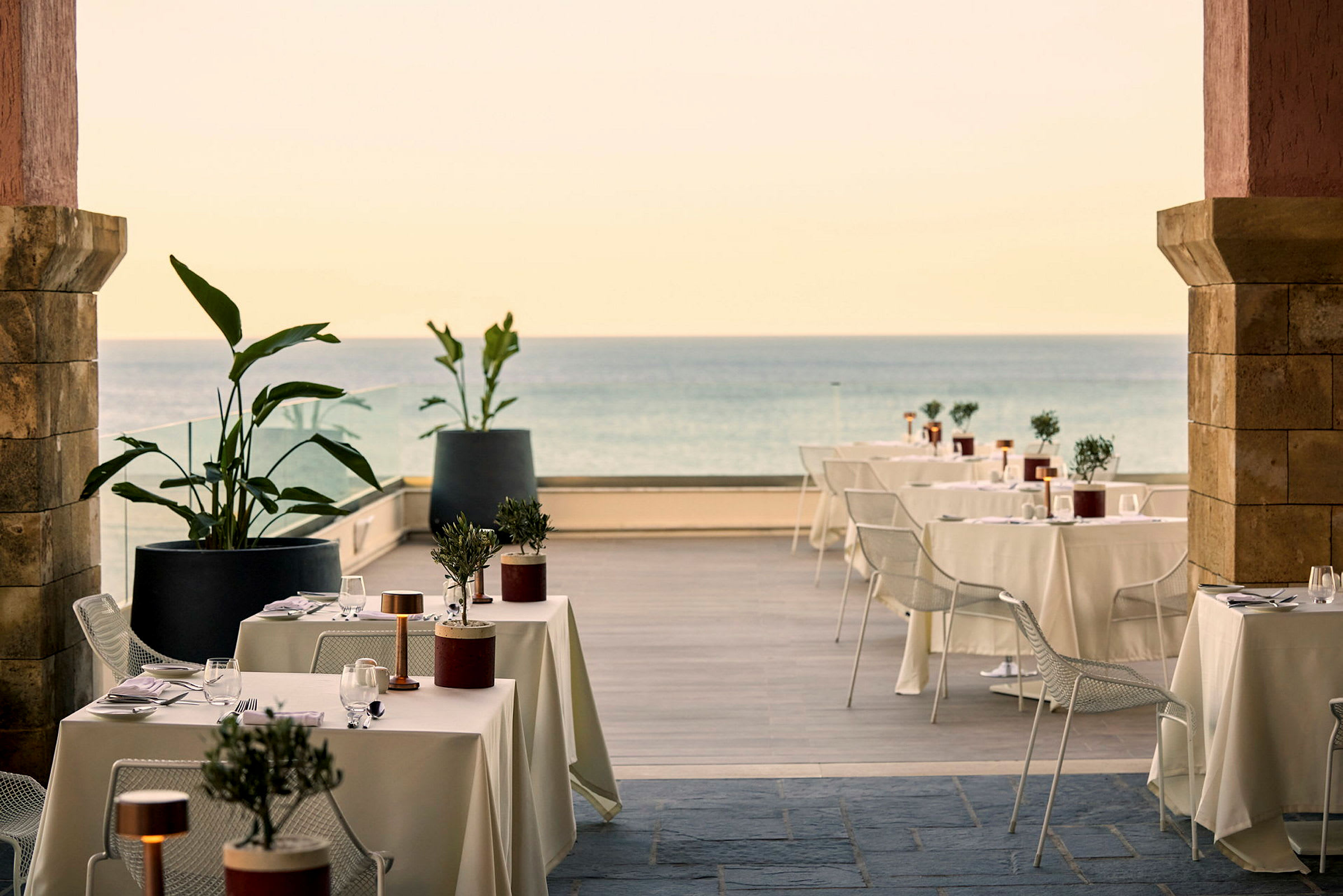 Utendørsrestaurant med dekkede bord, stoler og dekorative planter nær havet. (AI-generert tekst)