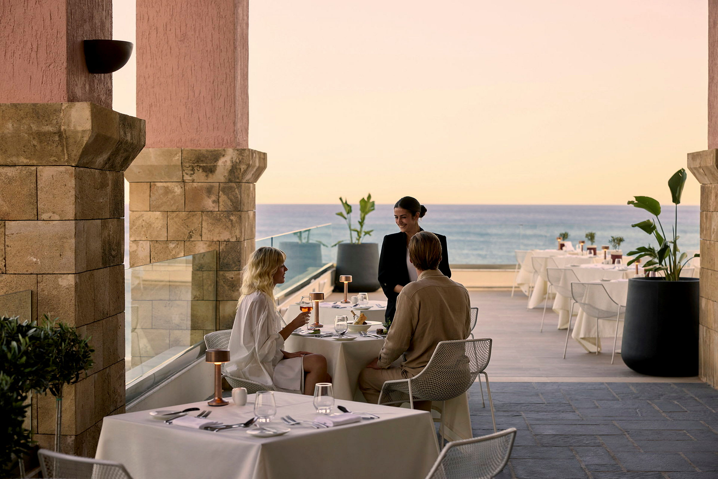 Personer som sitter ved et dekket restaurantbord med utsikt over havet. (AI-generert tekst)