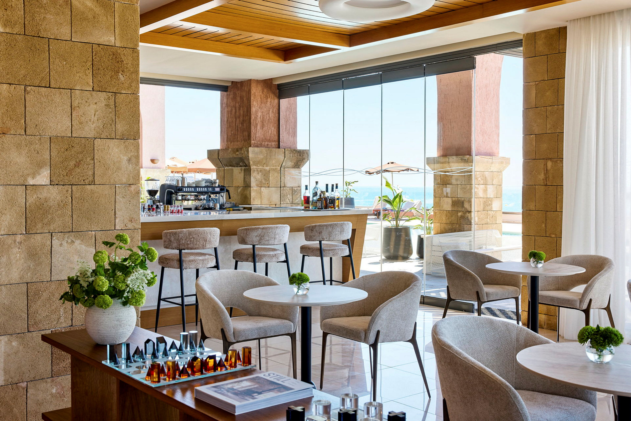 Bilde av en moderne og lys lounge med sittegrupper, bord og dekorative detaljer. Vinduer gir utsikt mot havet. (AI-generert tekst)
