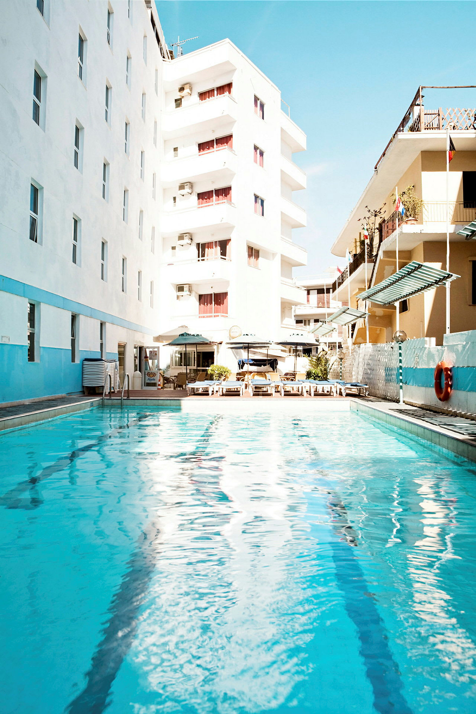 Et utendørsbasseng omgitt av hotellbygninger og solsenger. (AI-generert tekst)