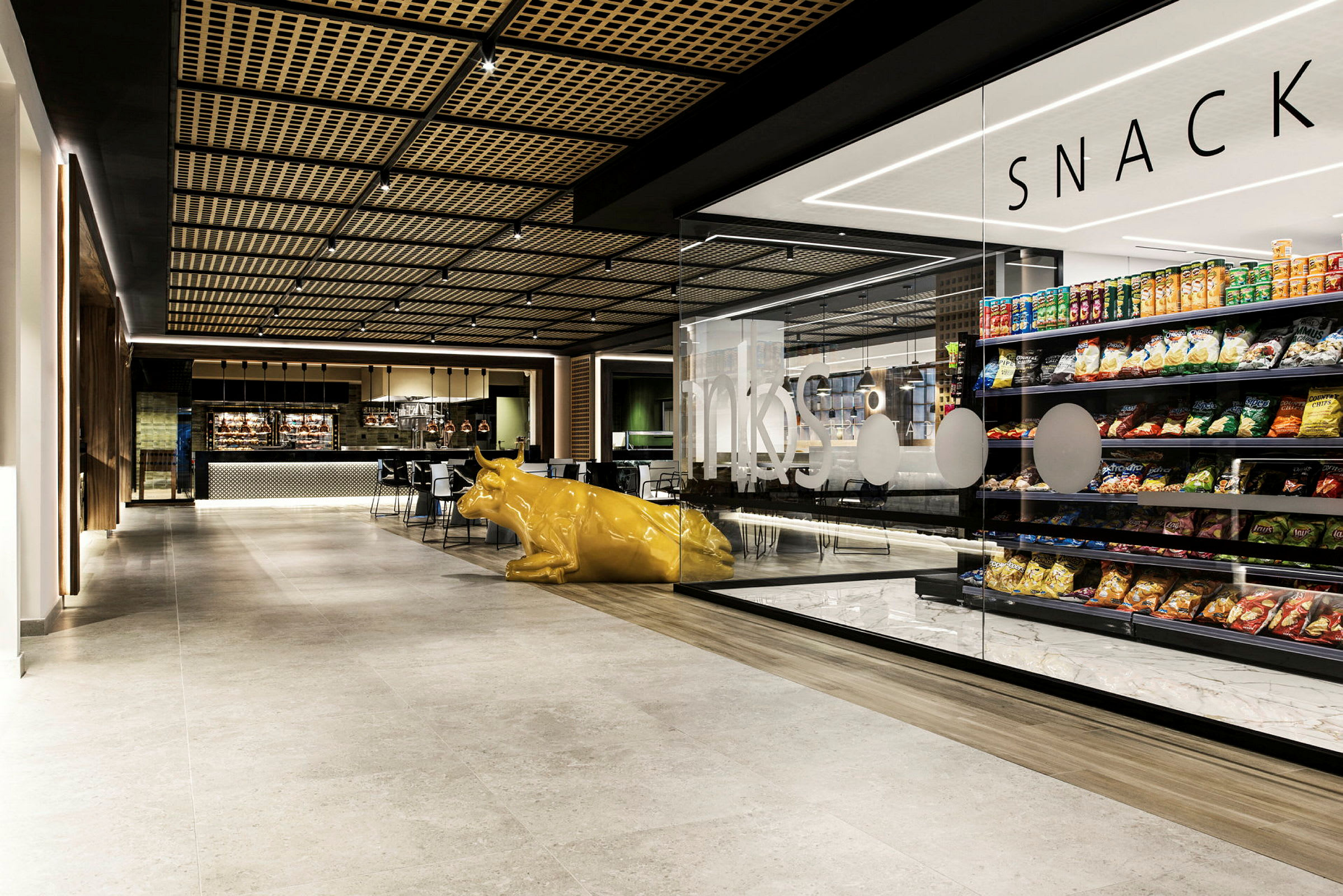 Et innendørsbilde av en moderne kafé med butikkhyller fylt med snacks og en gul kufigur i forgrunnen. (AI-generert tekst)