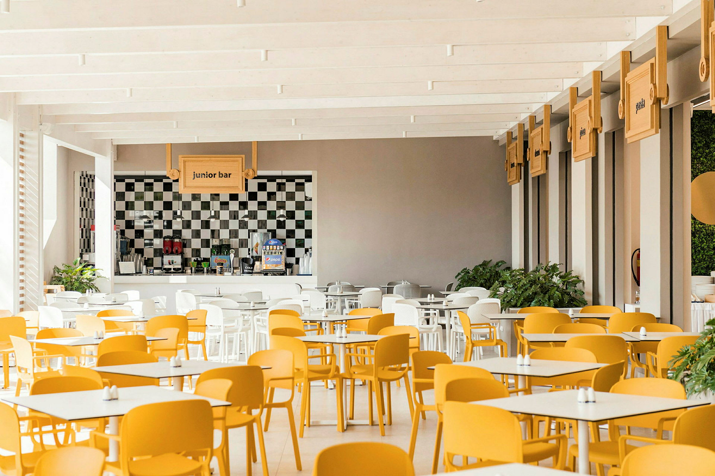 En lys spisesal med gule og hvite stoler og bordoppsett. En bardisk merket "junior bar" er synlig i bakgrunnen. (AI-generert tekst)