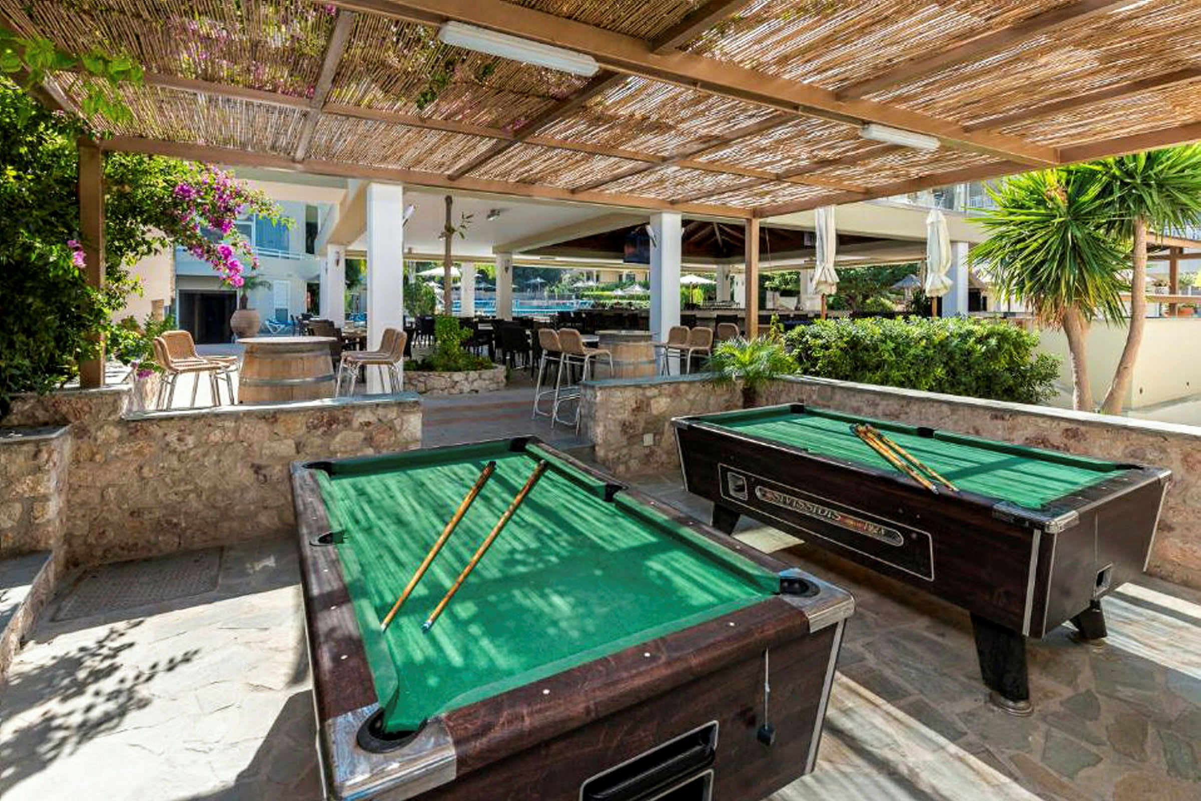 Grønne billiardbord under en pergola av tre, omgitt av planter og sitteplasser. (AI-generert tekst)