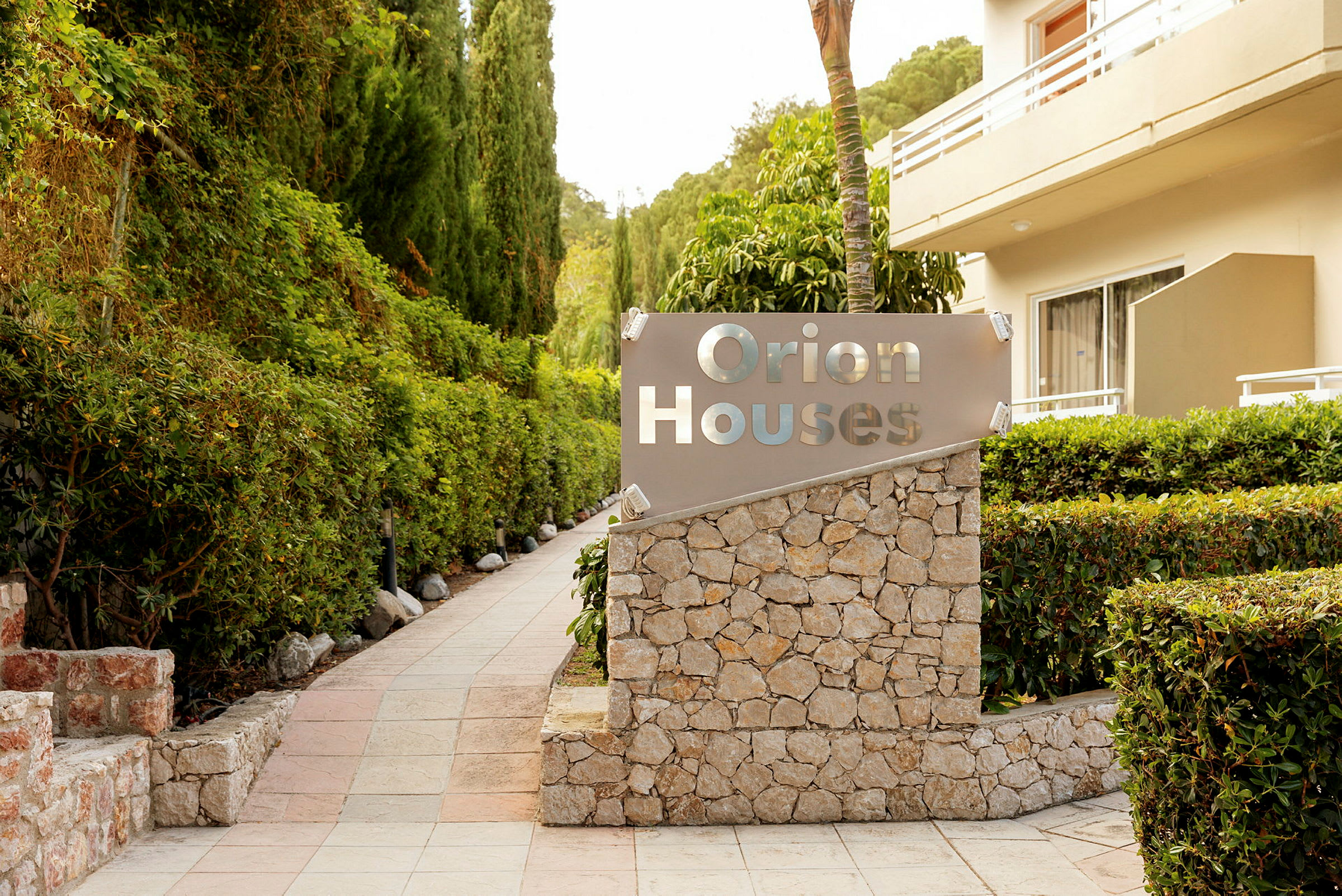 Skilt med teksten 'Orion Houses' ved siden av en gangvei omgitt av grøntområder og en bygning. (AI-generert tekst)