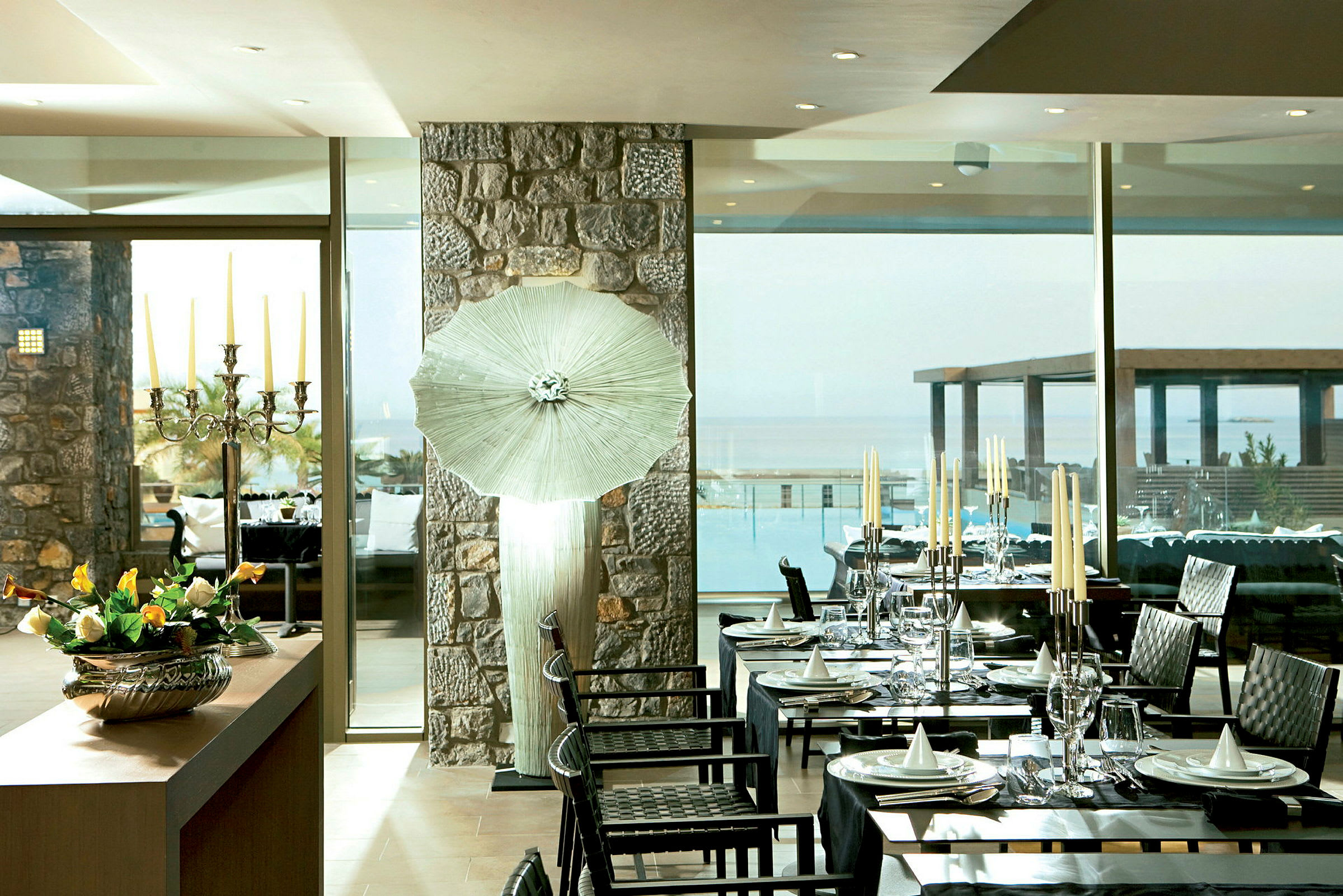 En innendørsrestaurant med dekkede bord, en stor vindusvegg med utsikt over et basseng og havet samt en dekorert lysestake og blomster på et bord. (AI-generert tekst)