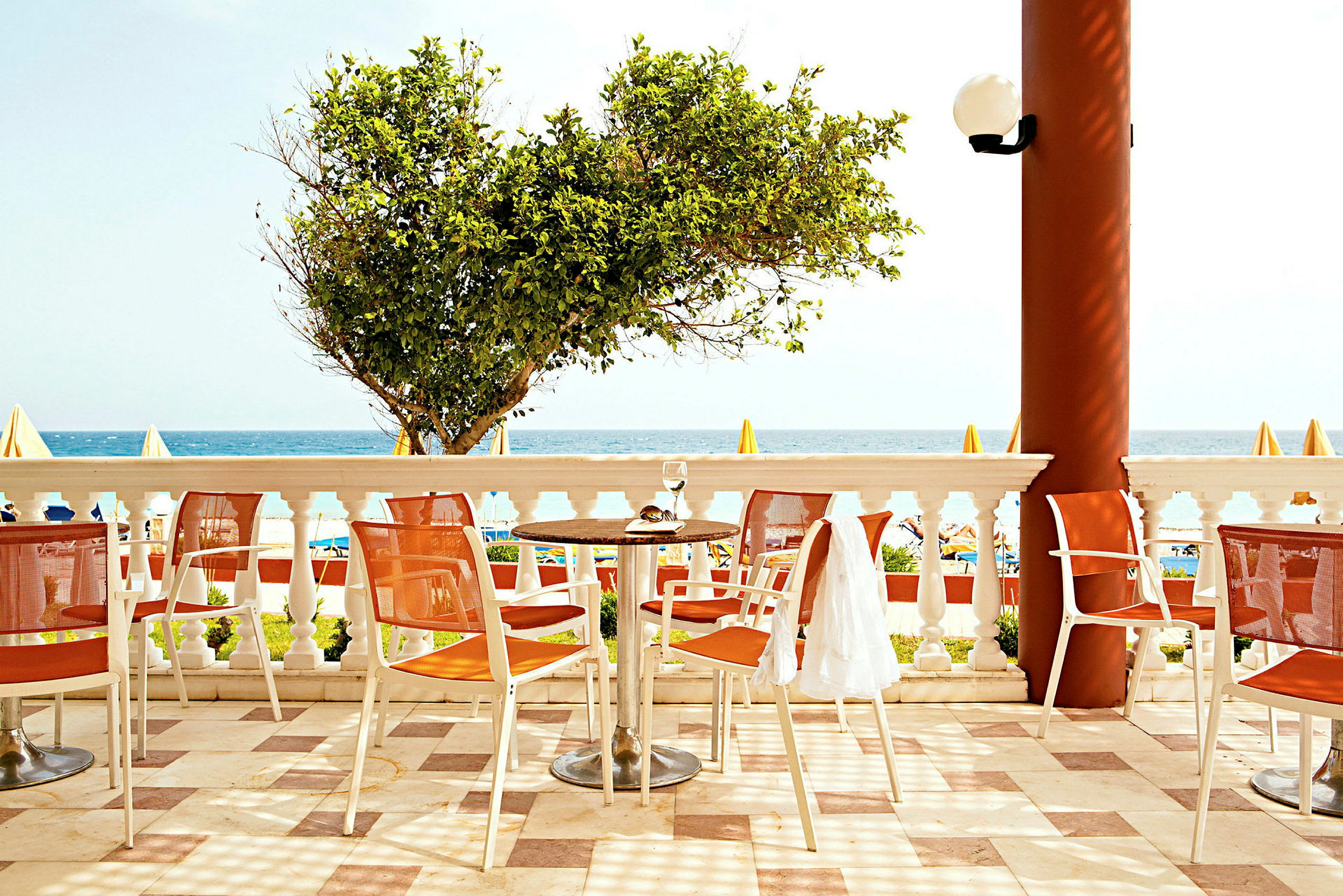 En terrasse med bord og stoler, havet i bakgrunnen og en hage med et tre. (AI-generert tekst)