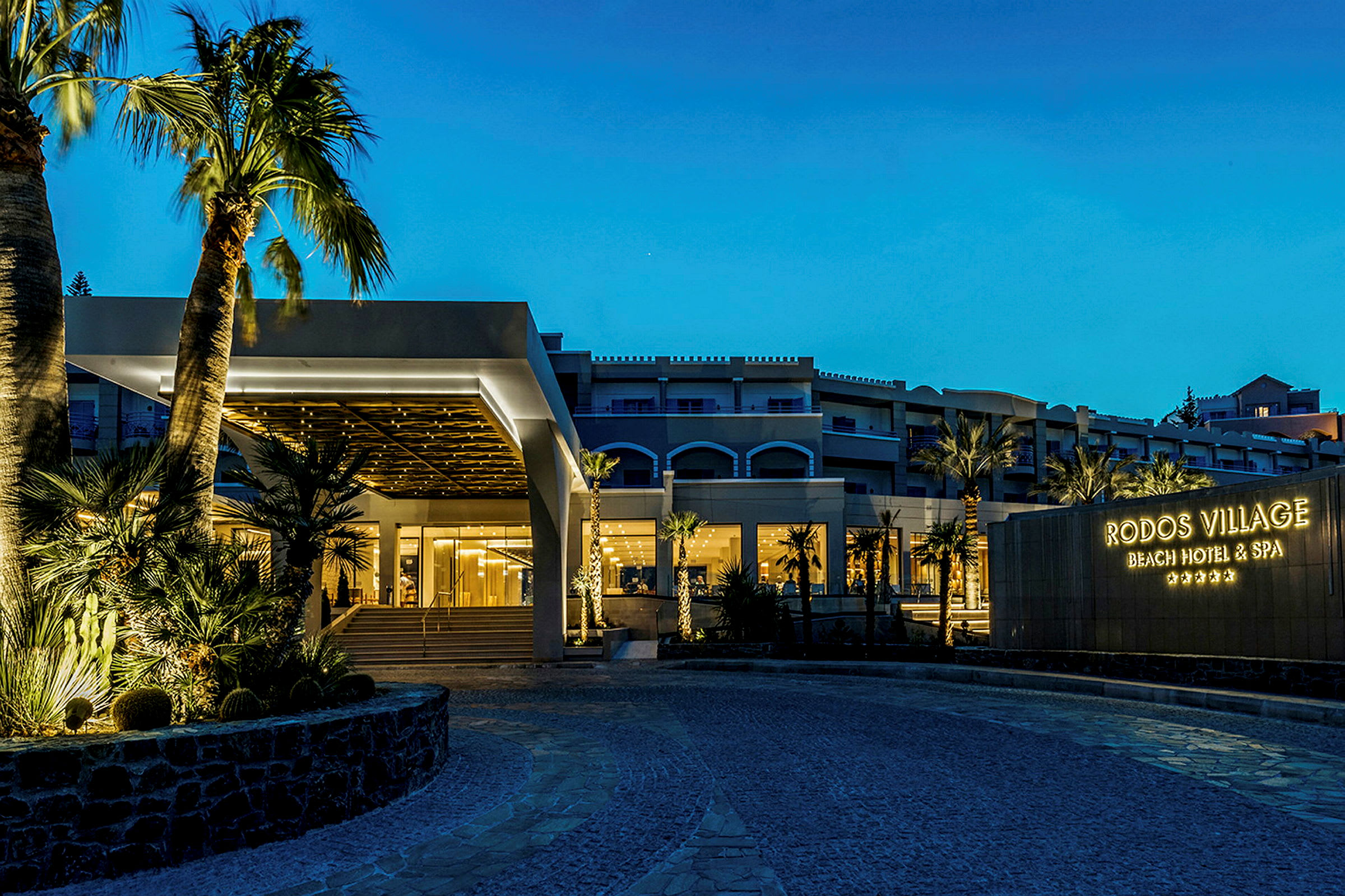 Et hotell om kvelden med opplyst inngang og skilt med teksten 'Rodos Village Beach Hotel & Spa'. (AI-generert tekst)