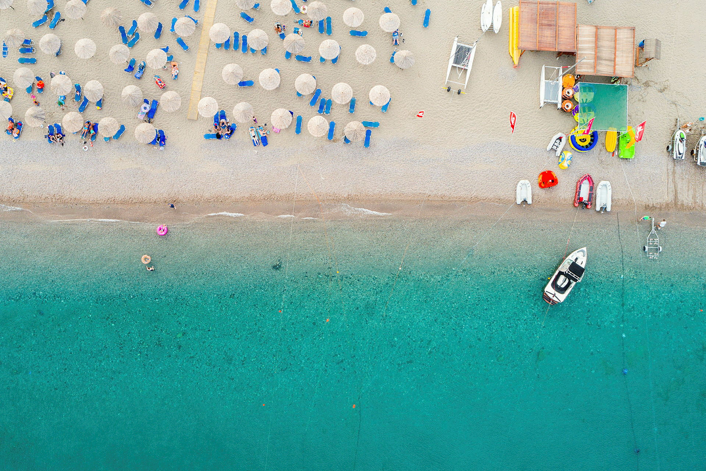 Flyfotografi over en strand med solsenger, parasoller og aktiviteter. Nær stranden vises klart turkist vann og en båt. (AI-generert tekst)