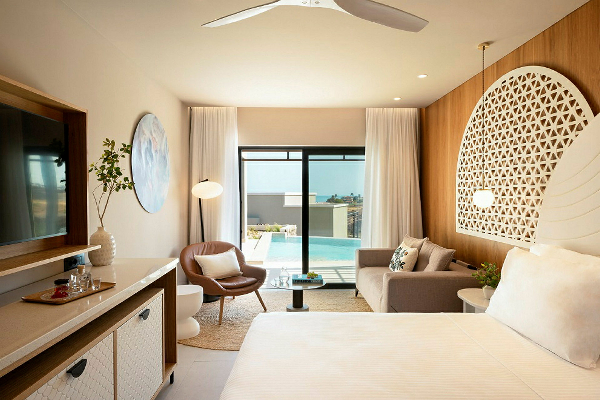 Et elegant hotellrom med moderne møbler, en sofa, en lenestol, et sengområde med dekorativt hodegjerde, et stort speil og glassdører som åpner ut mot et svømmebasseng utendørs. (AI-generert tekst)