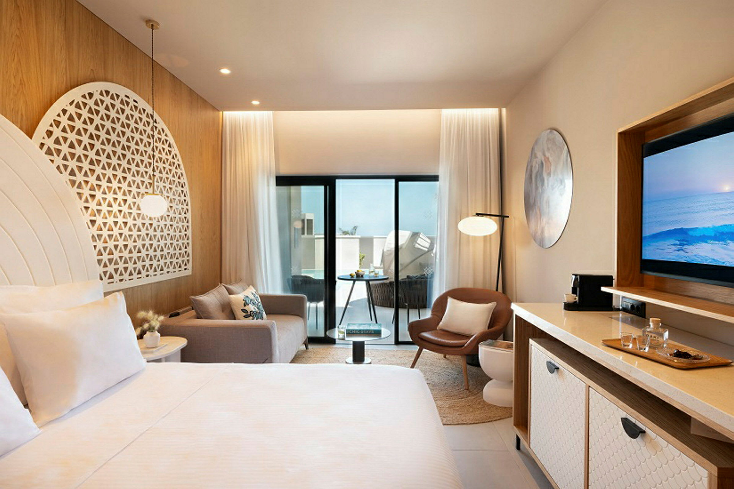 Et moderne hotellrom med lyse møbler og dekorasjoner, inkludert en seng, sofa, stol og TV. Rommet har store glassdører som fører ut til en balkong. (AI-generert tekst)