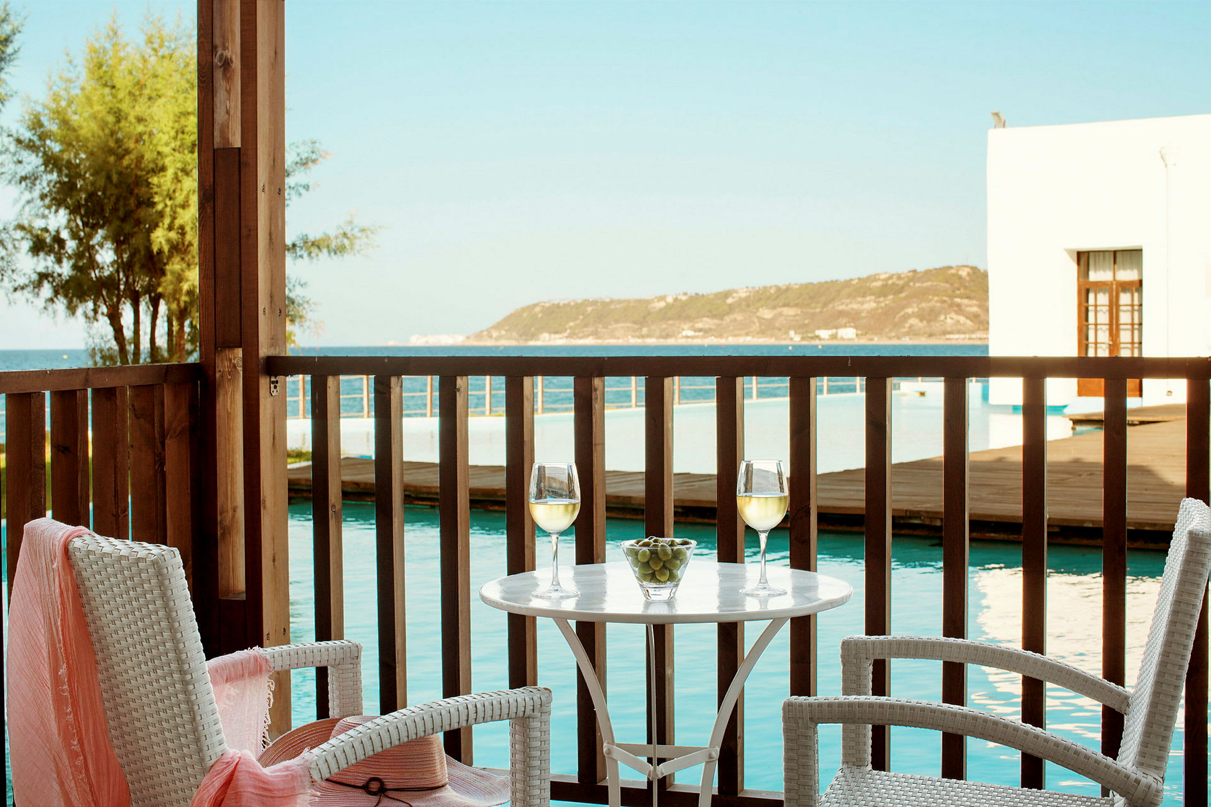 To glass hvitvin og en skål med druer på et bord på en terrasse med utsikt mot havet. (AI-generert tekst)