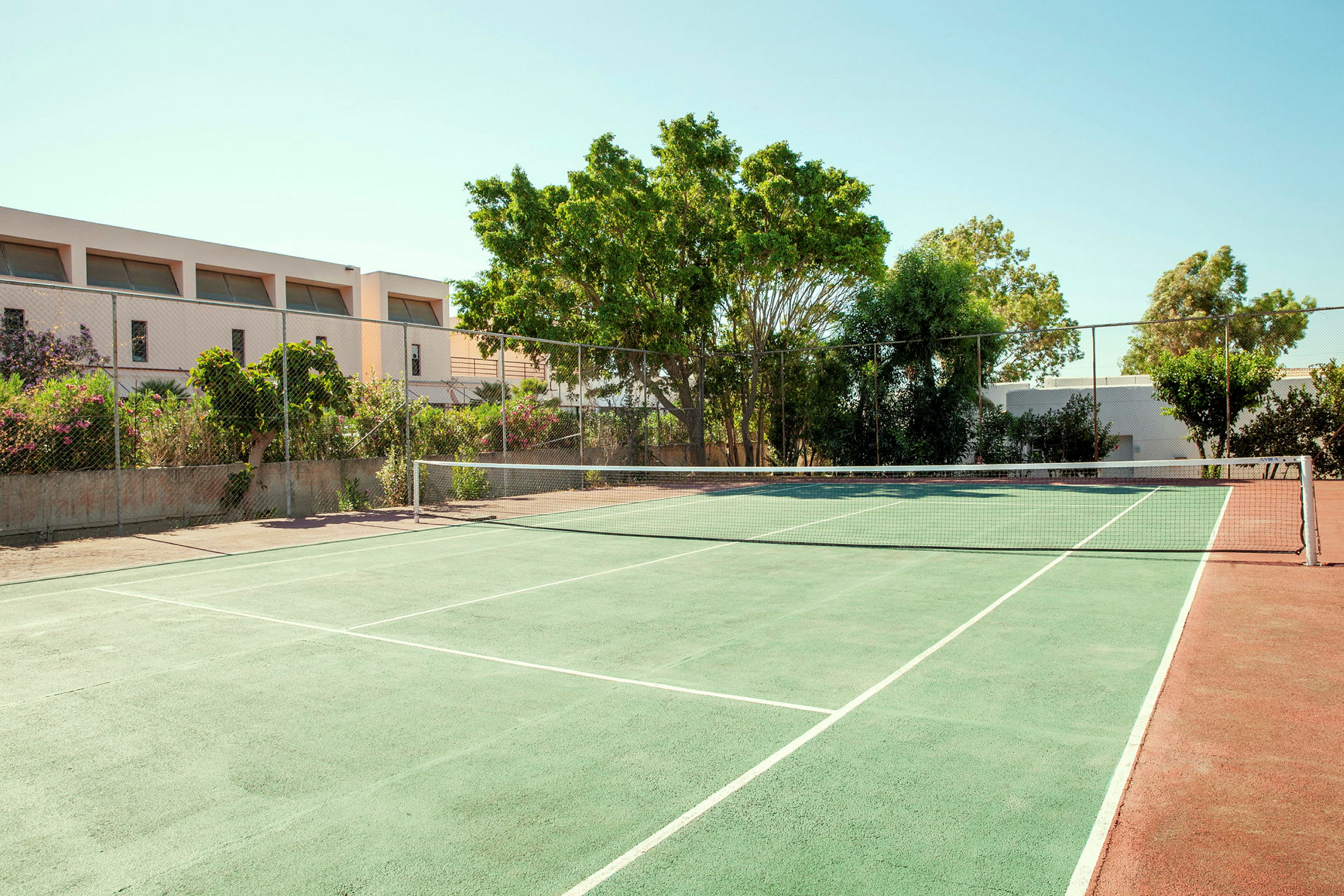 En tennisbane med grønn overflate og nett, omgitt av trær og bygninger. (AI-generert tekst)