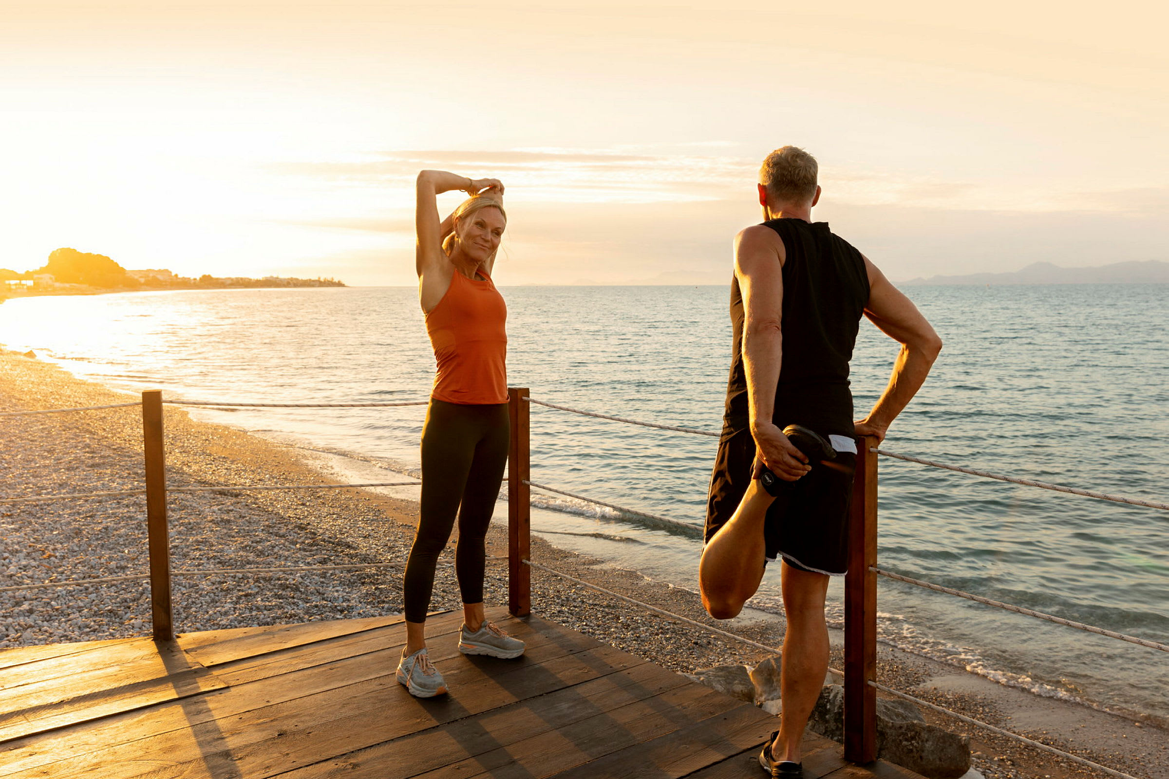 To personer strekker ut på en trebrygge ved en strand ved solnedgang. (AI-generert tekst)