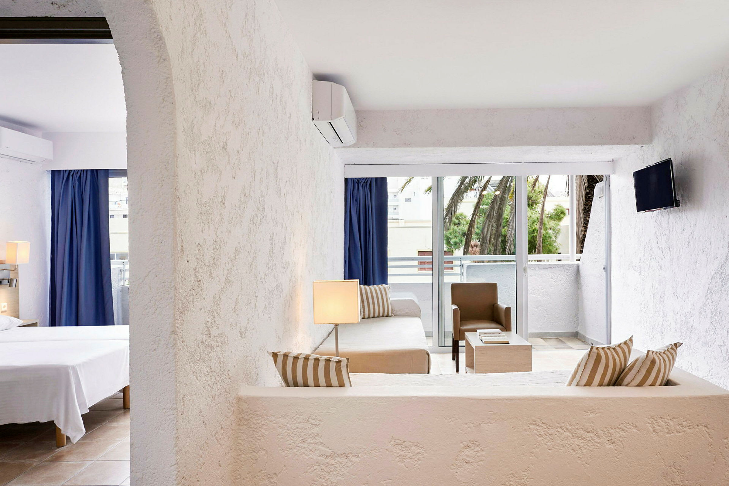 Et lyst hotellrom med en stuedel, møbler som sofa, bord og stol i nøytrale farger, og et åpent soverom. Det er en balkong med utsikt mot palmer. (AI-generert tekst)