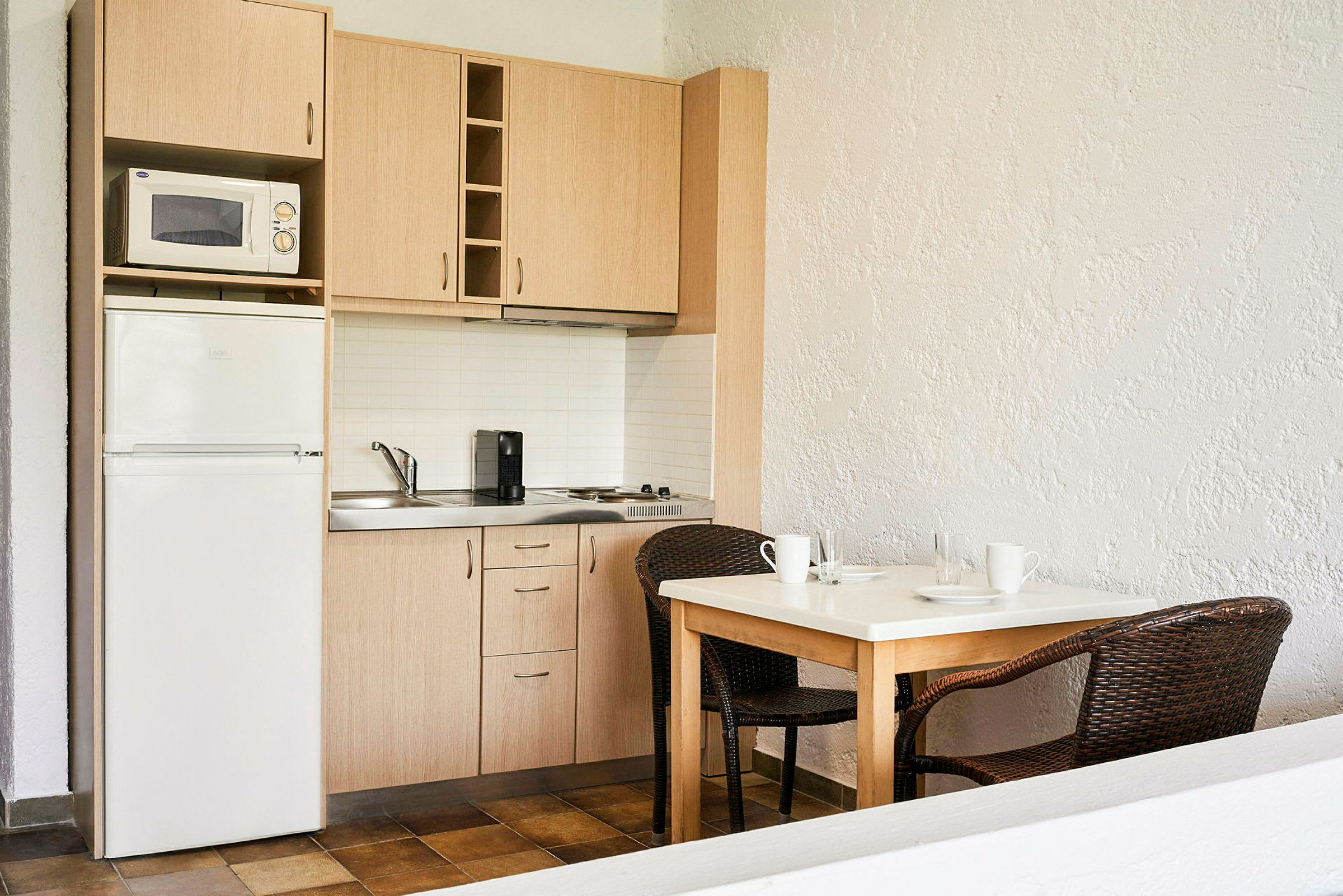 Et kompakt kjøkken med treskap, en mikrobølgeovn, et kjøleskap og en kaffemaskin. Foran kjøkkenet står et bord med to rottingstoler. (AI-generert tekst)