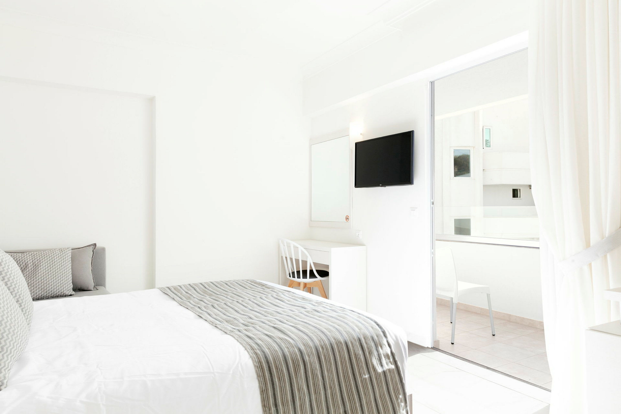 Interiør av et lyst hotellrom med en seng og skrivebord ved siden av en hvit stol. En TV er montert på veggen, og glassdører fører til en balkong. (AI-generert tekst)