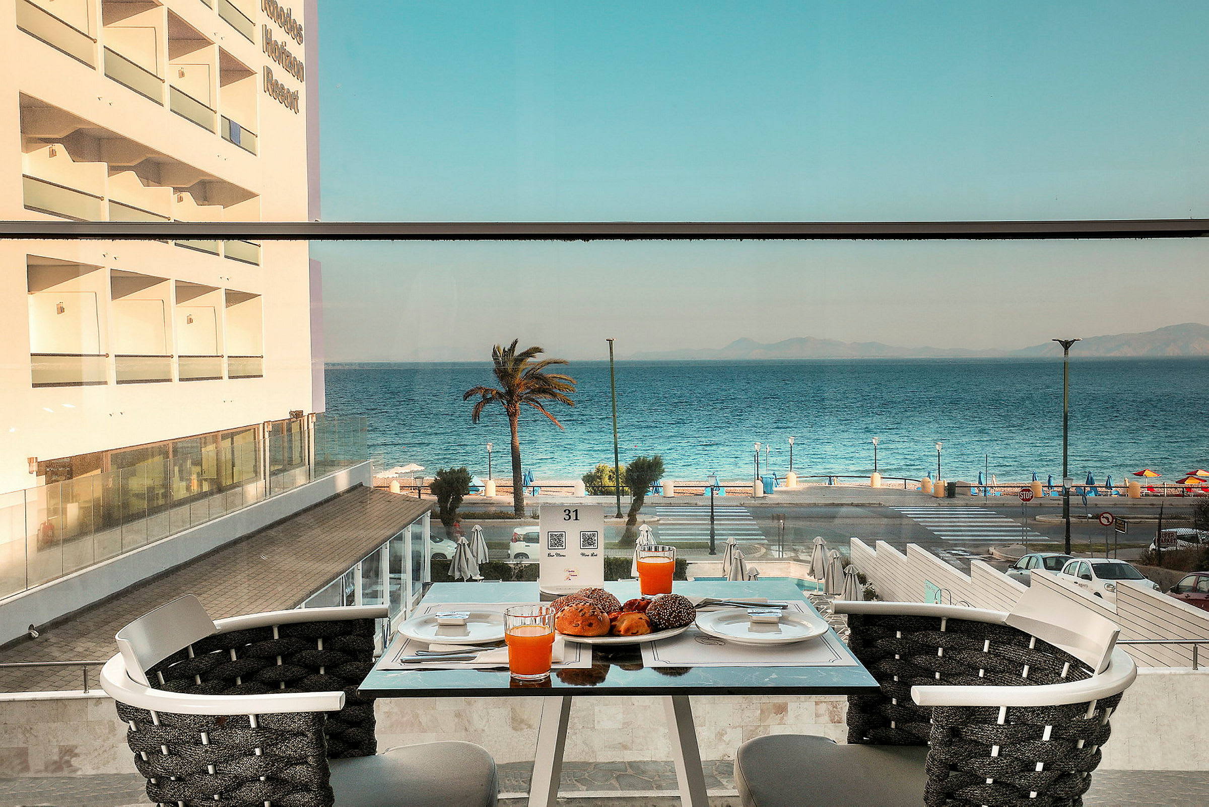 Utsikt fra et bord på en terrasse med appelsinjuice og bakverk, mot havet og en palme. (AI-generert tekst)