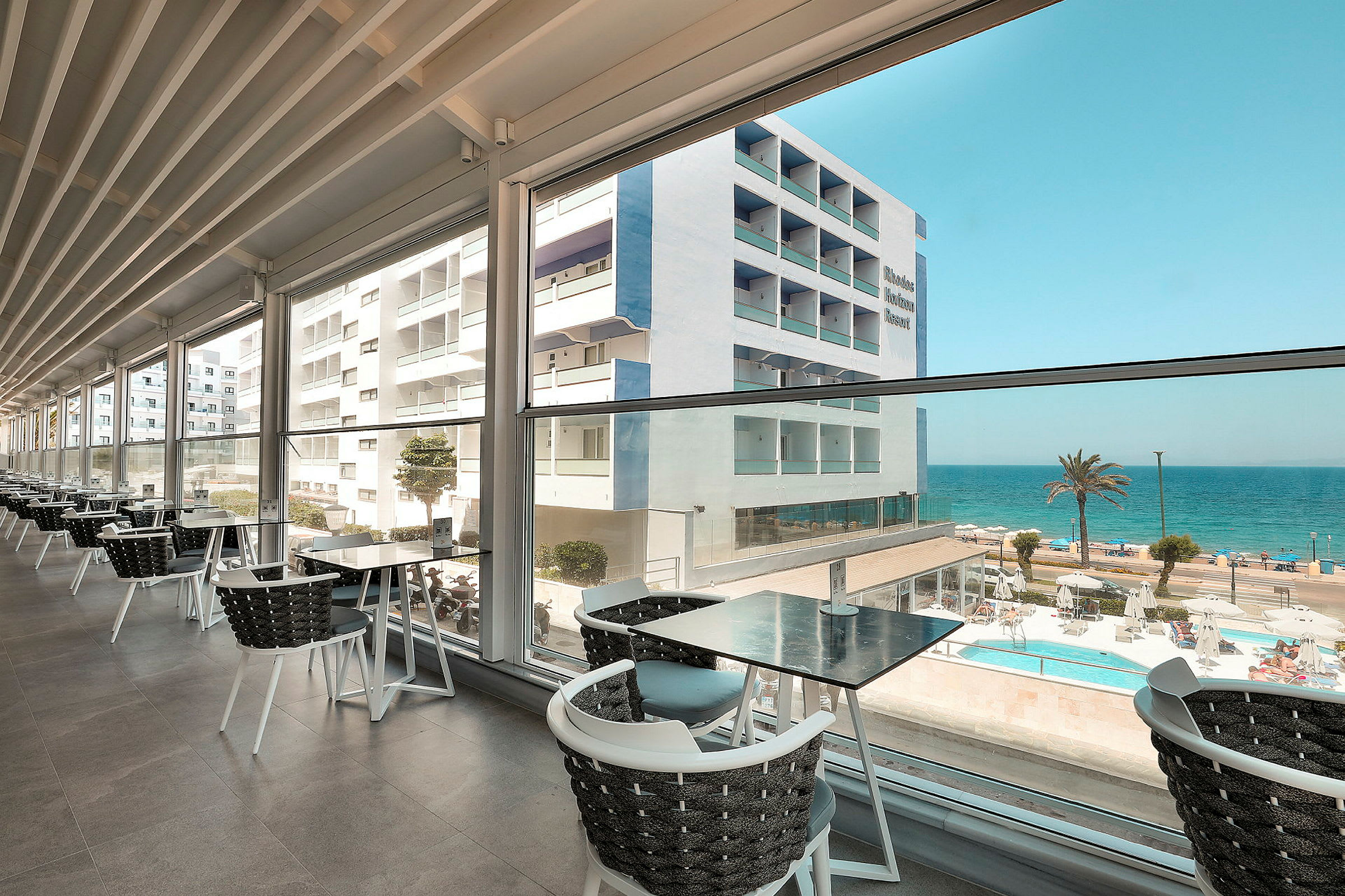 Utsikt fra en restaurant med glassvegger mot en bygning, et basseng og havet. (AI-generert tekst)