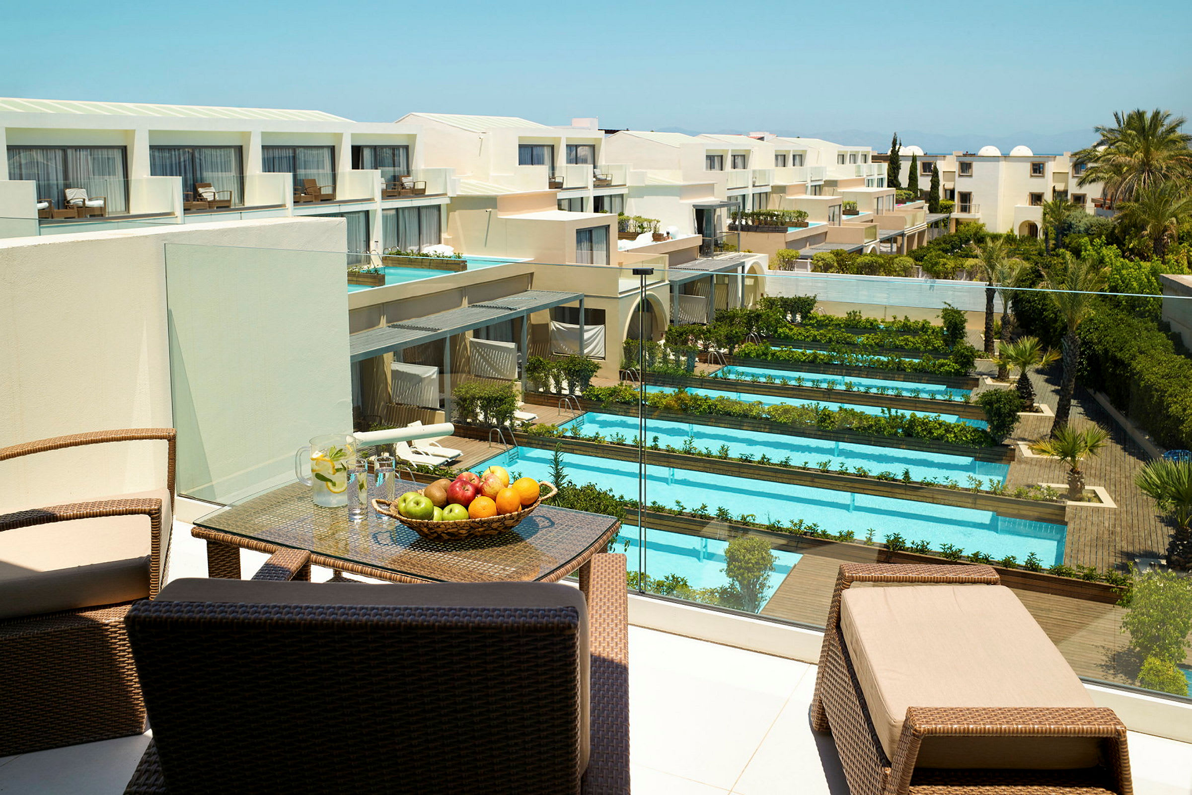 Hotellområde med flere bassenger og grøntområder, samt en terrasse med møbler og frukt. Utsikt over en moderne bygning. (AI-generert tekst)