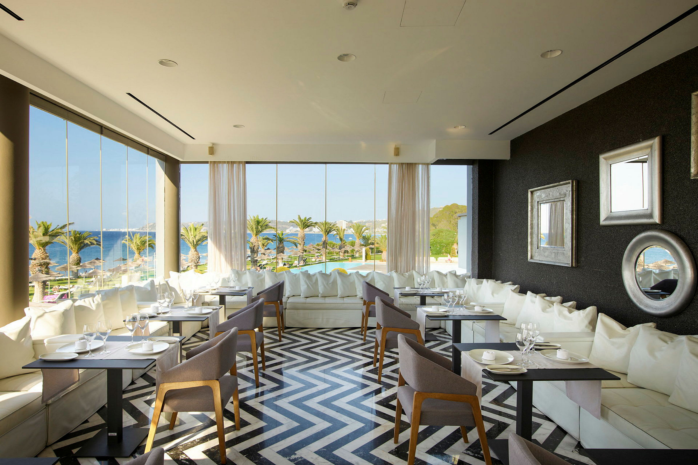 Interiør av en restaurant med store vinduer, moderne møbler og utsikt over palmer og havet. (AI-generert tekst)