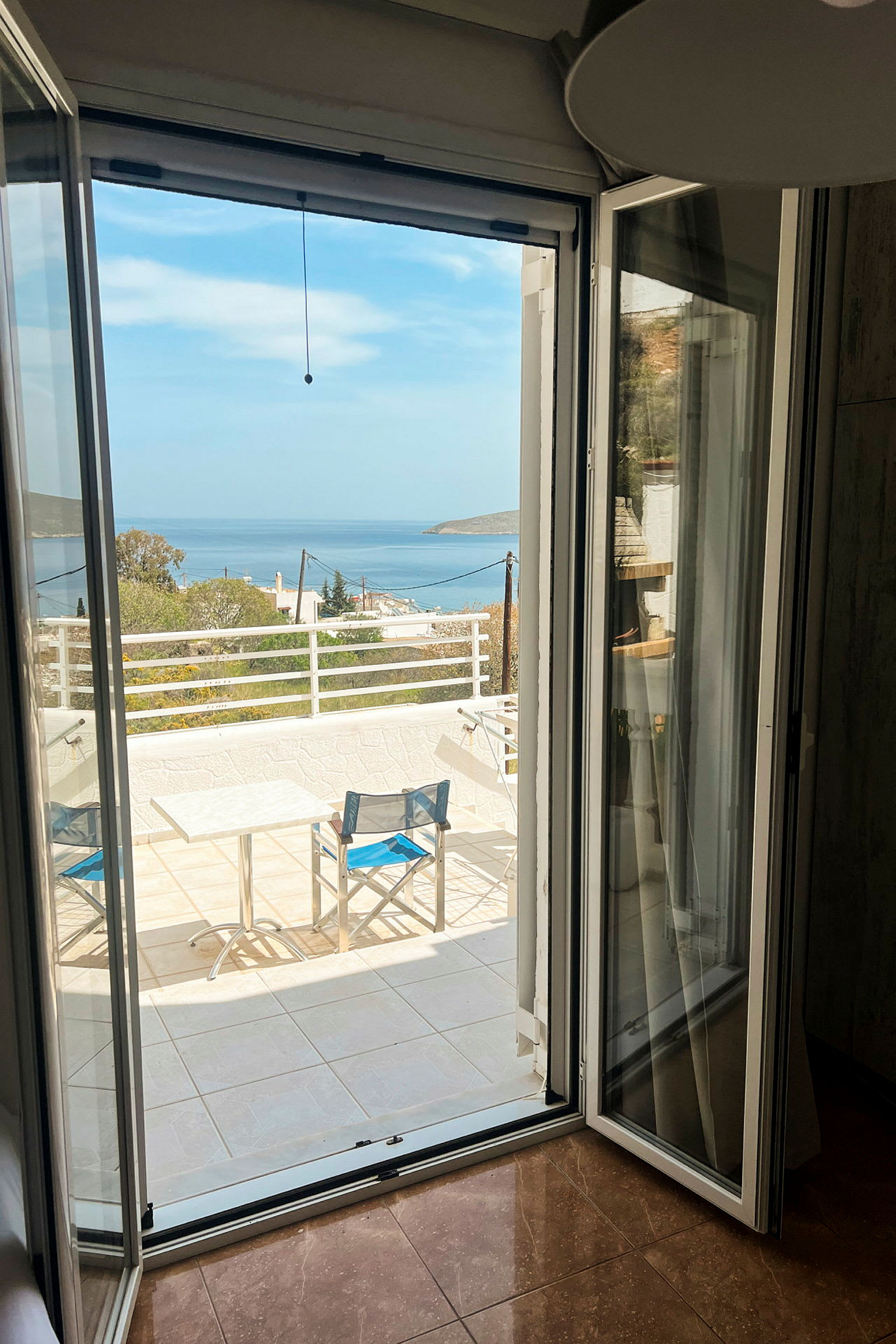Utsikt fra et åpent vindu mot en terrasse med bord og stoler, og havet i bakgrunnen. (AI-generert tekst)