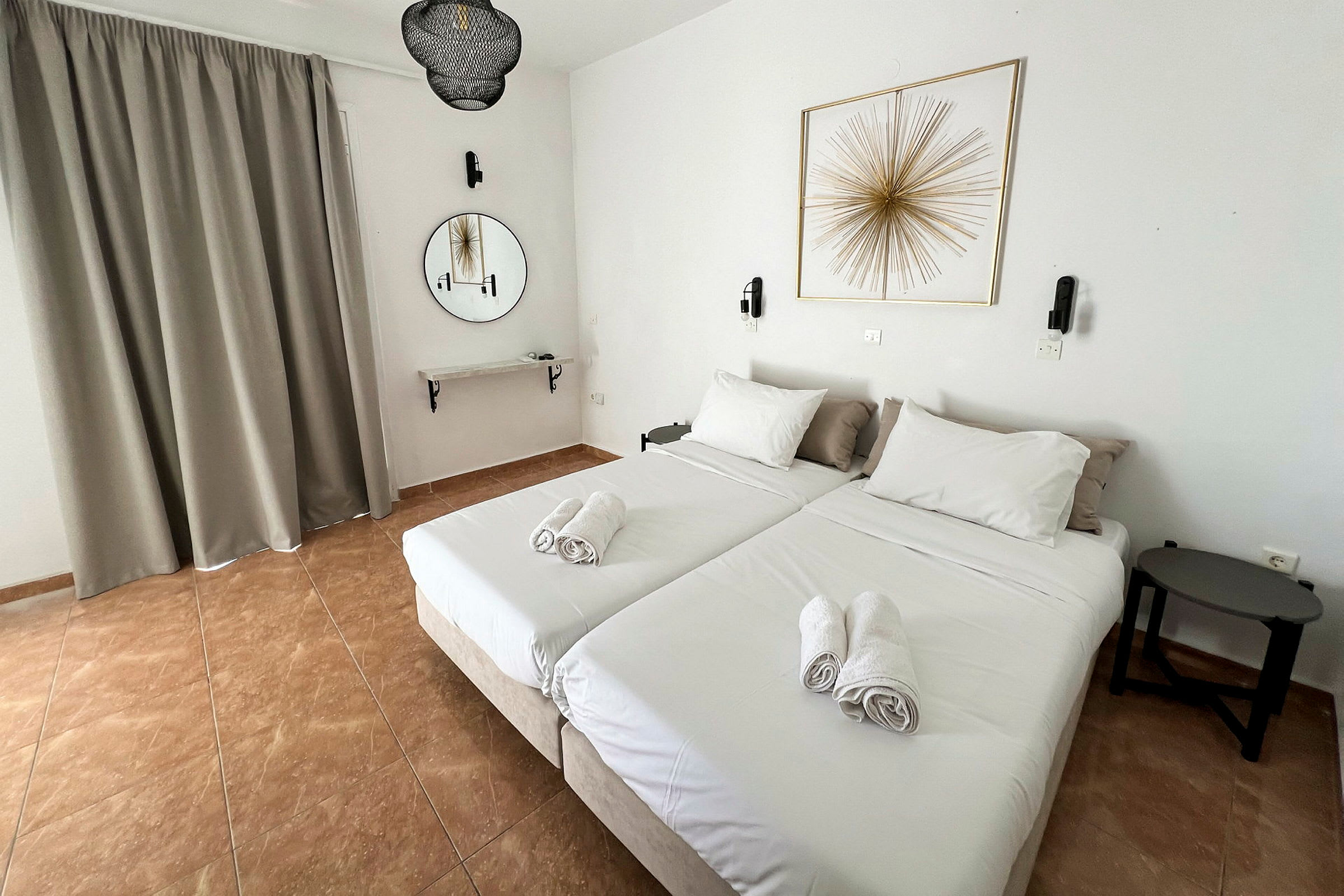 To enkeltsenger med hvitt sengetøy og brettede håndklær oppå står i et hotellrom. Et rundt speil og en veggdekorasjon ses på veggen. (AI-generert tekst)