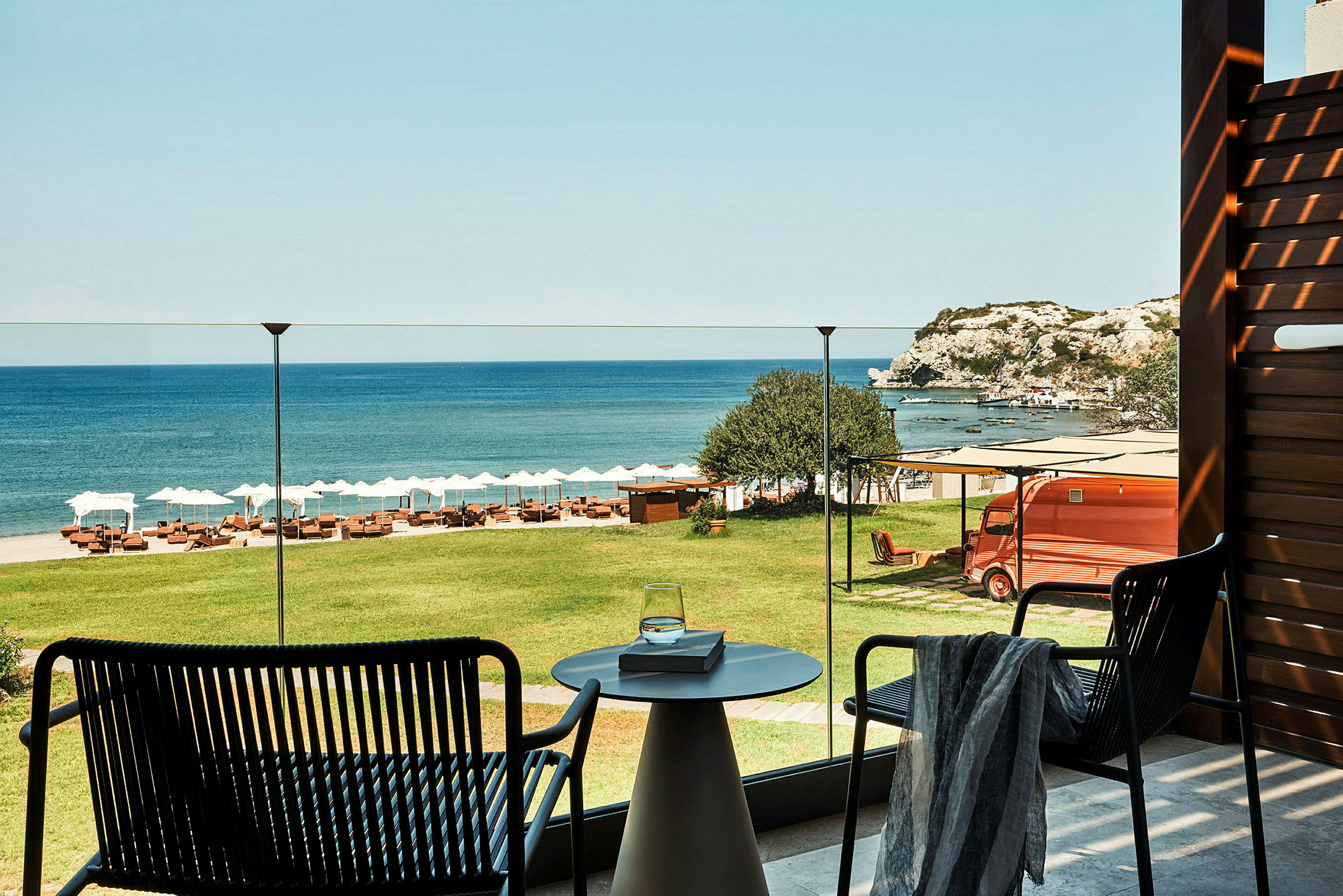 Utsikt fra en terrasse over grønt gress, en strand med solsenger og parasoller samt havet og klipper i bakgrunnen. (AI-generert tekst)