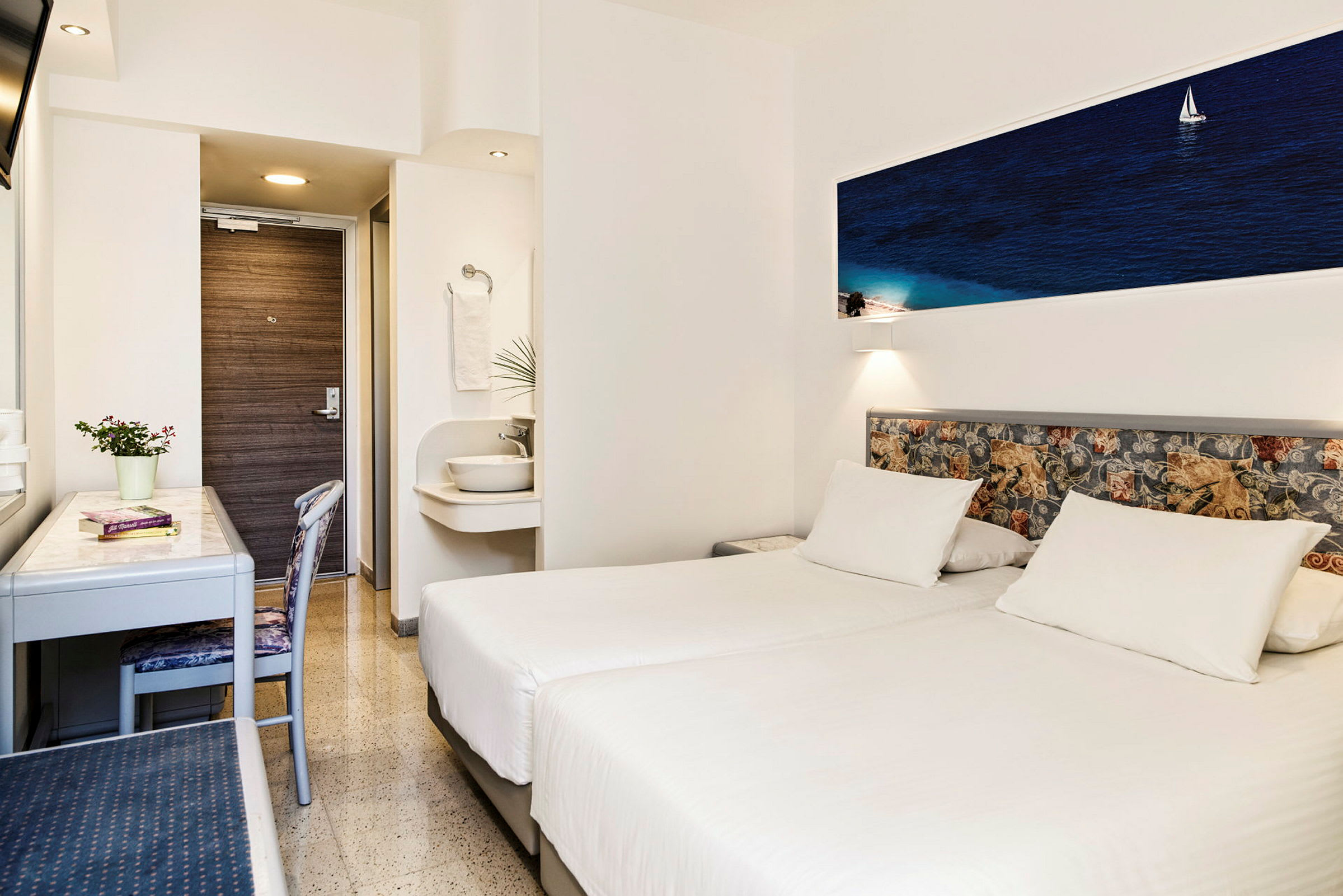 Et hotellrom med to hvite enkeltsenger, et skrivebord, en stol og en liten vask utenfor badet. (AI-generert tekst)