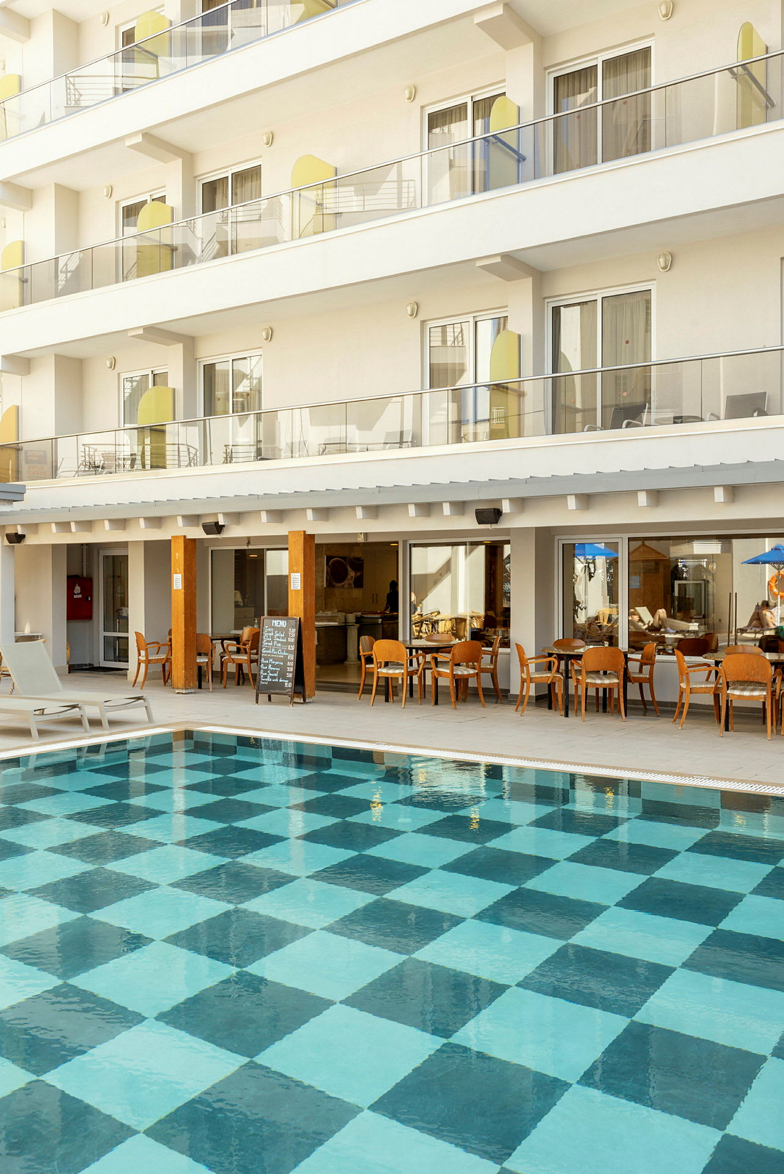Utendørsbasseng med rutet bunnmønster foran hotellbygning med balkonger og restaurant. (AI-generert tekst)