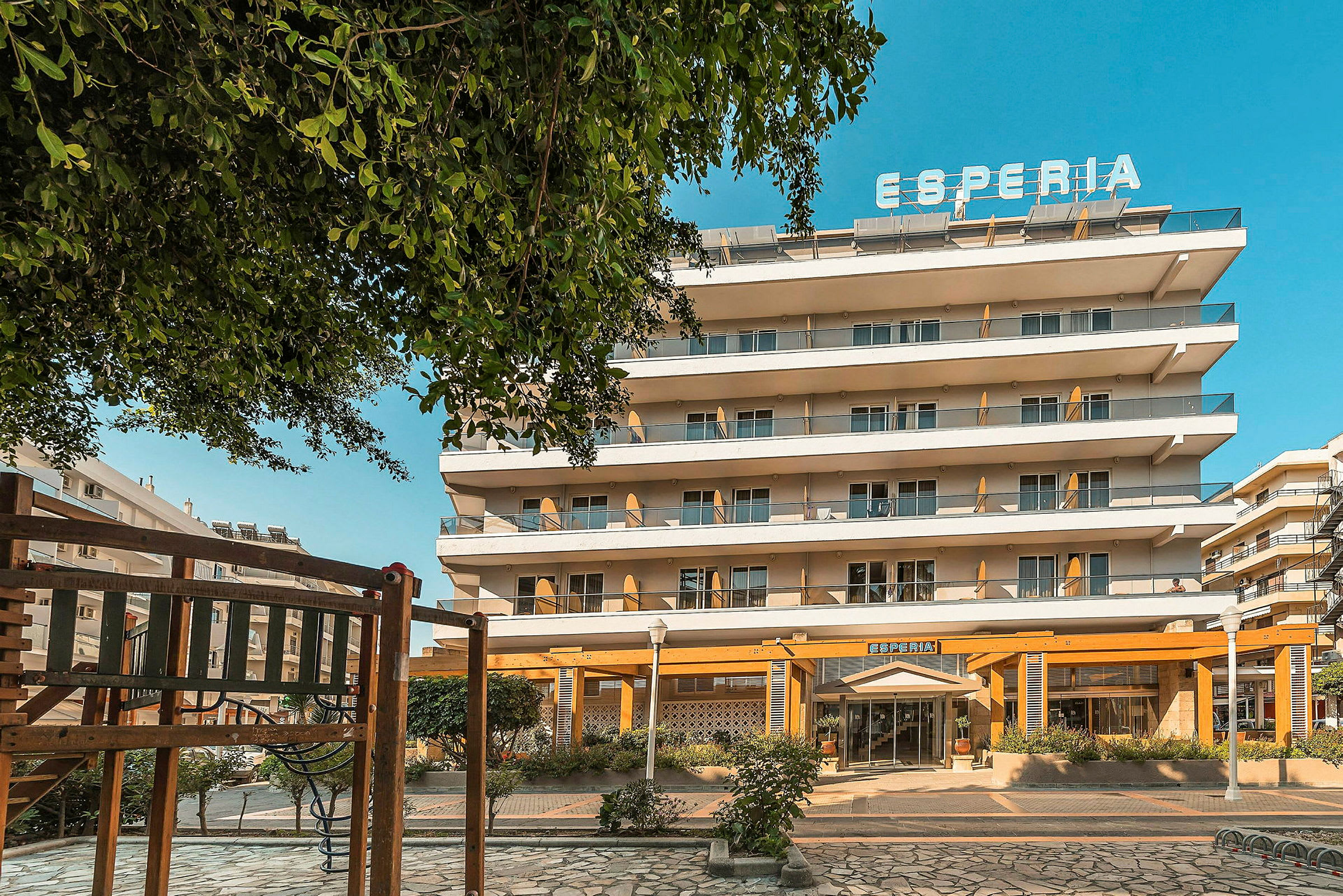 Hotellbygning med flere etasjer og et skilt med teksten 'Esperia' øverst. Det finnes lekeapparater i forgrunnen. (AI-generert tekst)