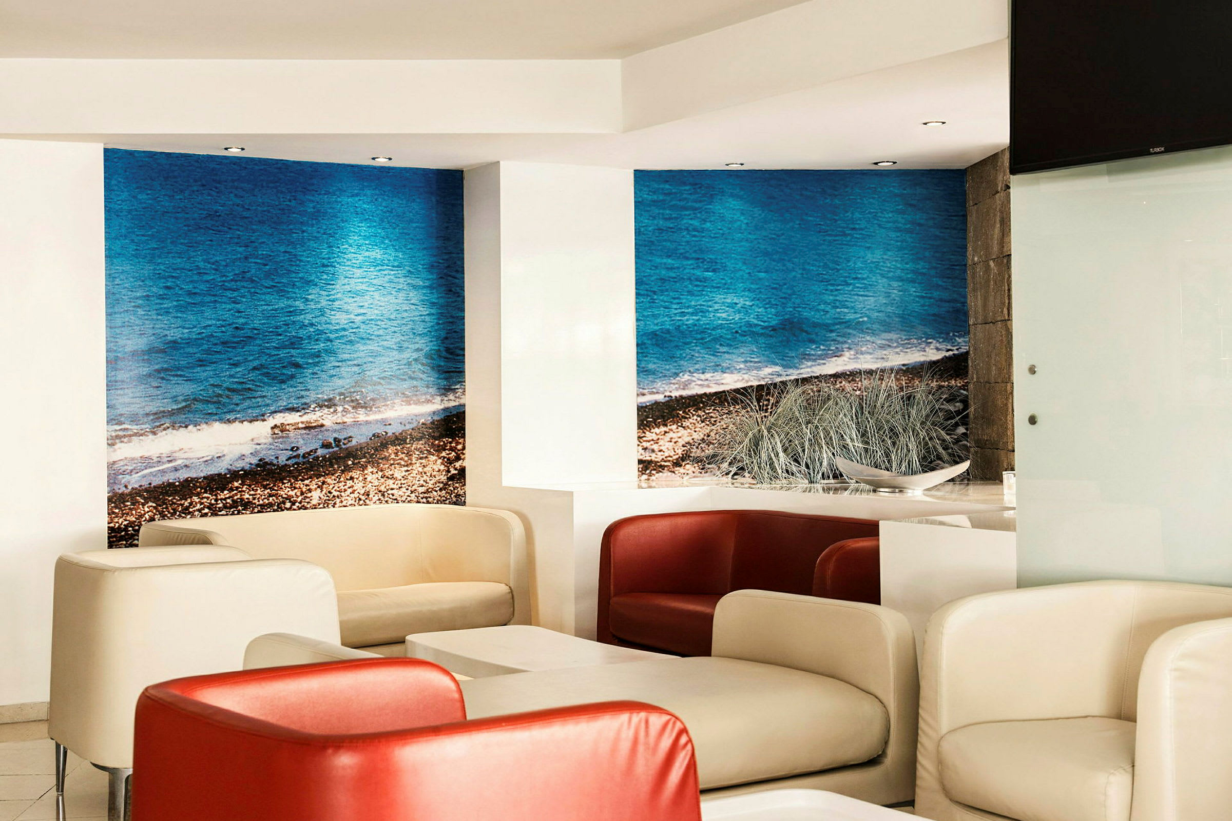 En moderne lounge med komfortable stoler i hvitt og rødt samt bord. Veggene har fototapet med motiv av en strand med blått hav. (AI-generert tekst)
