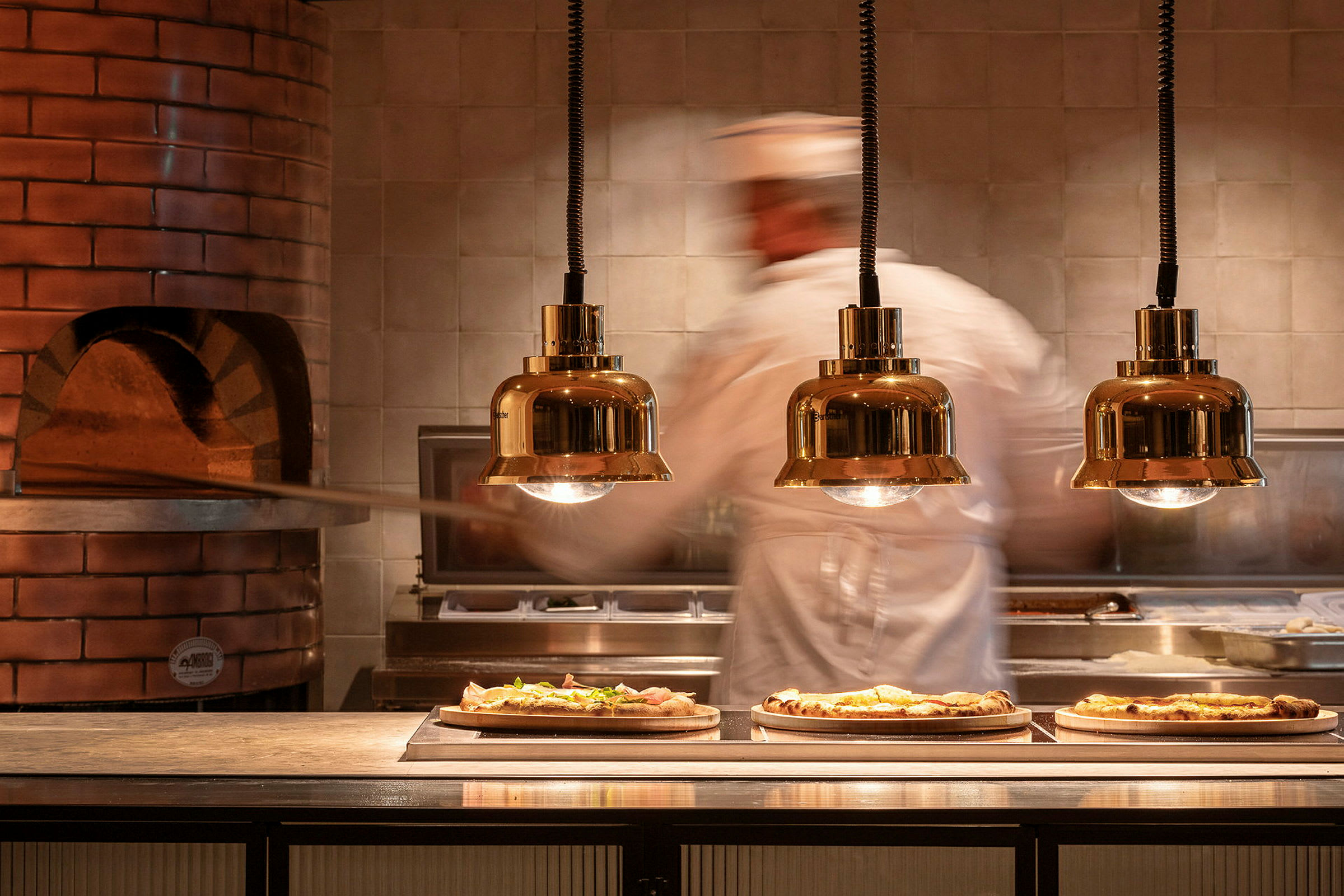 En pizzabaker jobber ved en ovn der tre ferdige pizzaer ligger på serveringsbordet. (AI-generert tekst)