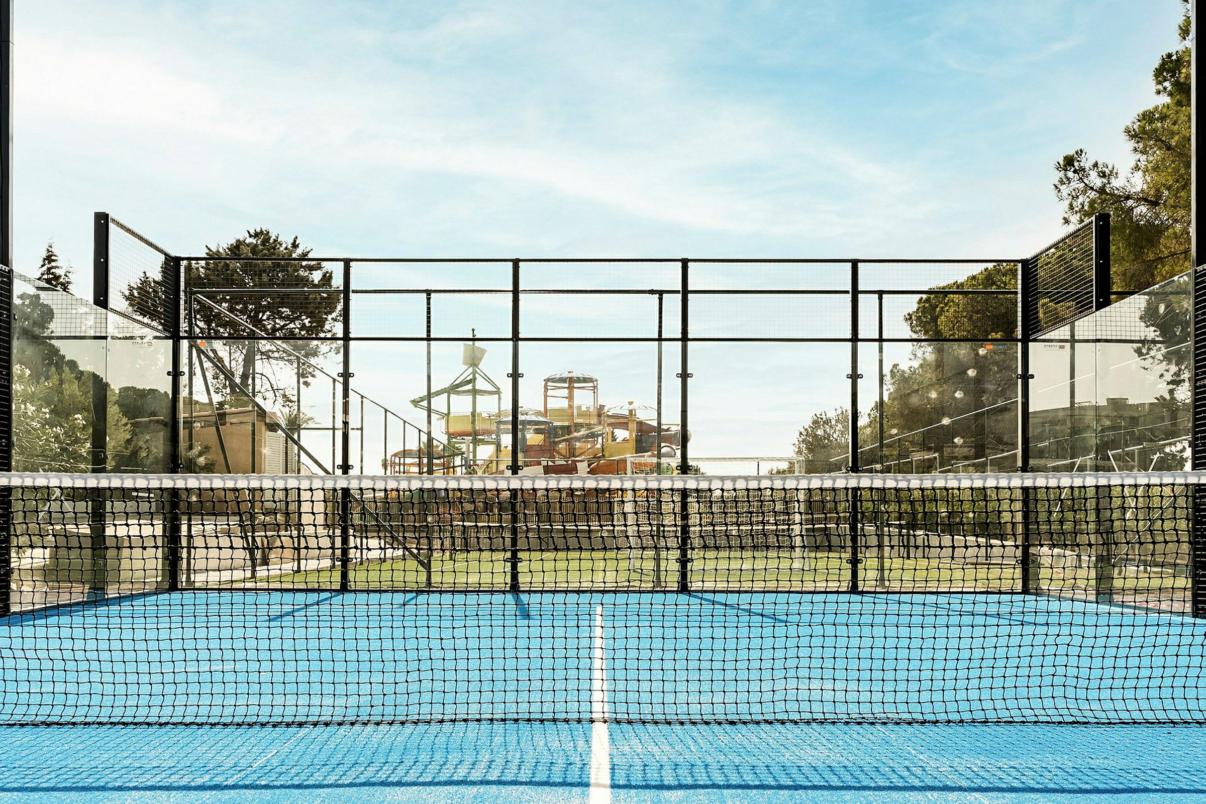 En padelbane med blått underlag og nett i forgrunnen, med en vannpark i bakgrunnen. (AI-generert tekst)