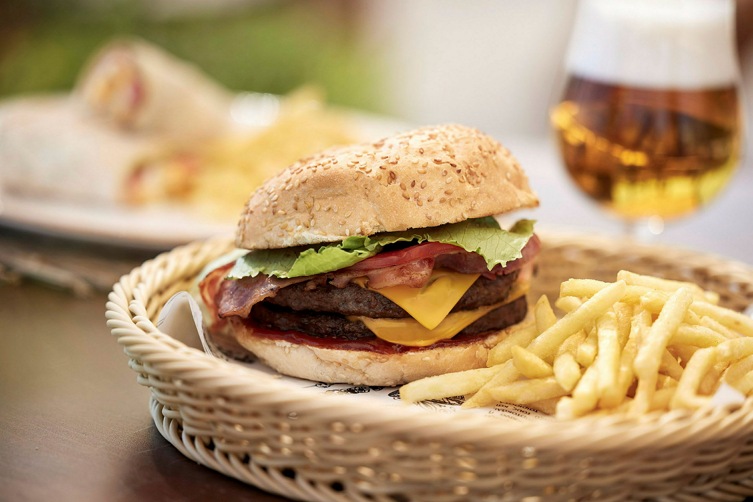 Hamburger med ost og bacon, servert med pommes frites i en kurv, samt en øl i bakgrunnen. (AI-generert tekst)
